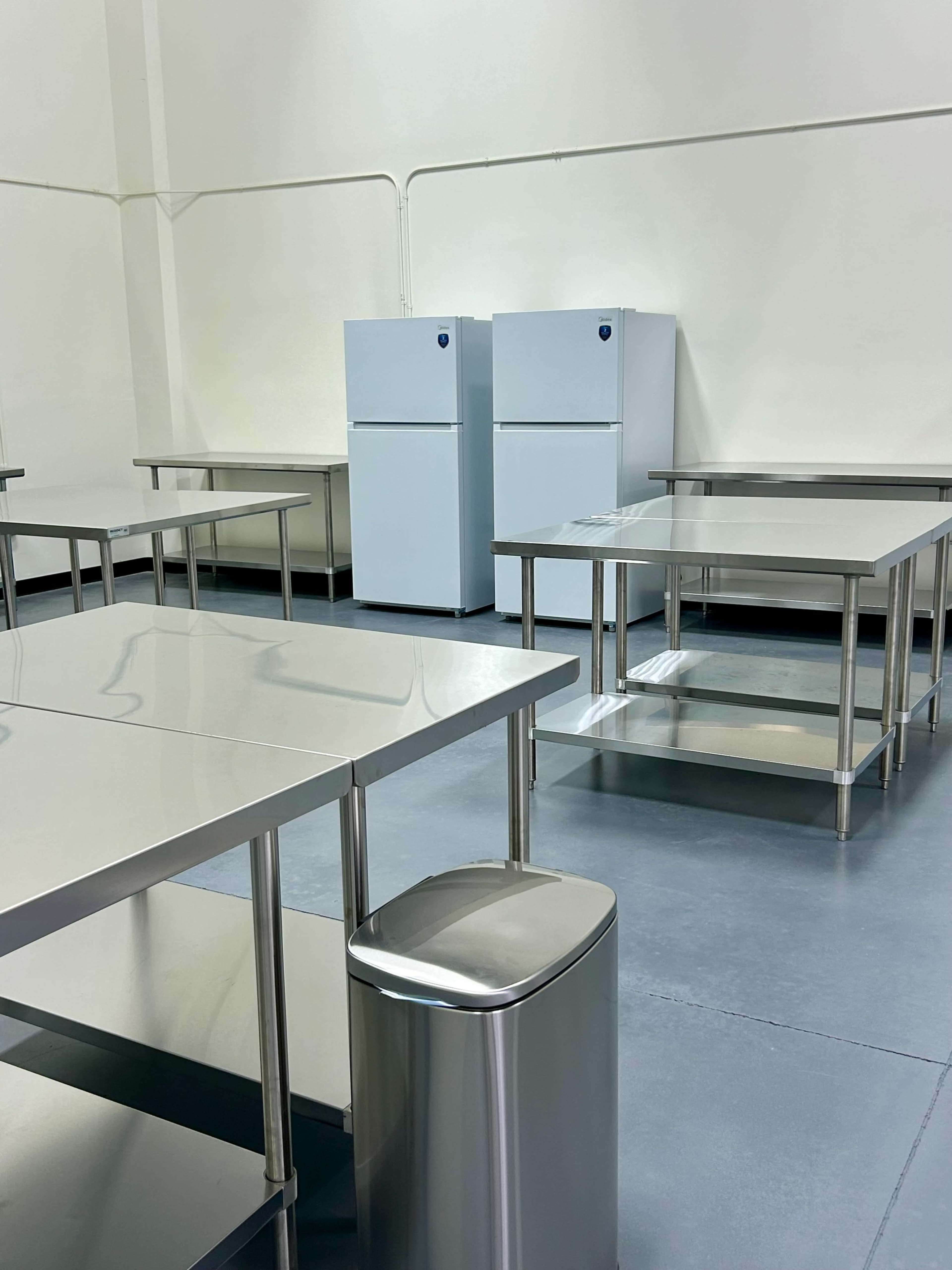 The image shows a spacious kitchen area with stainless steel tables, a trash bin, and two white refrigerators against a plain wall.