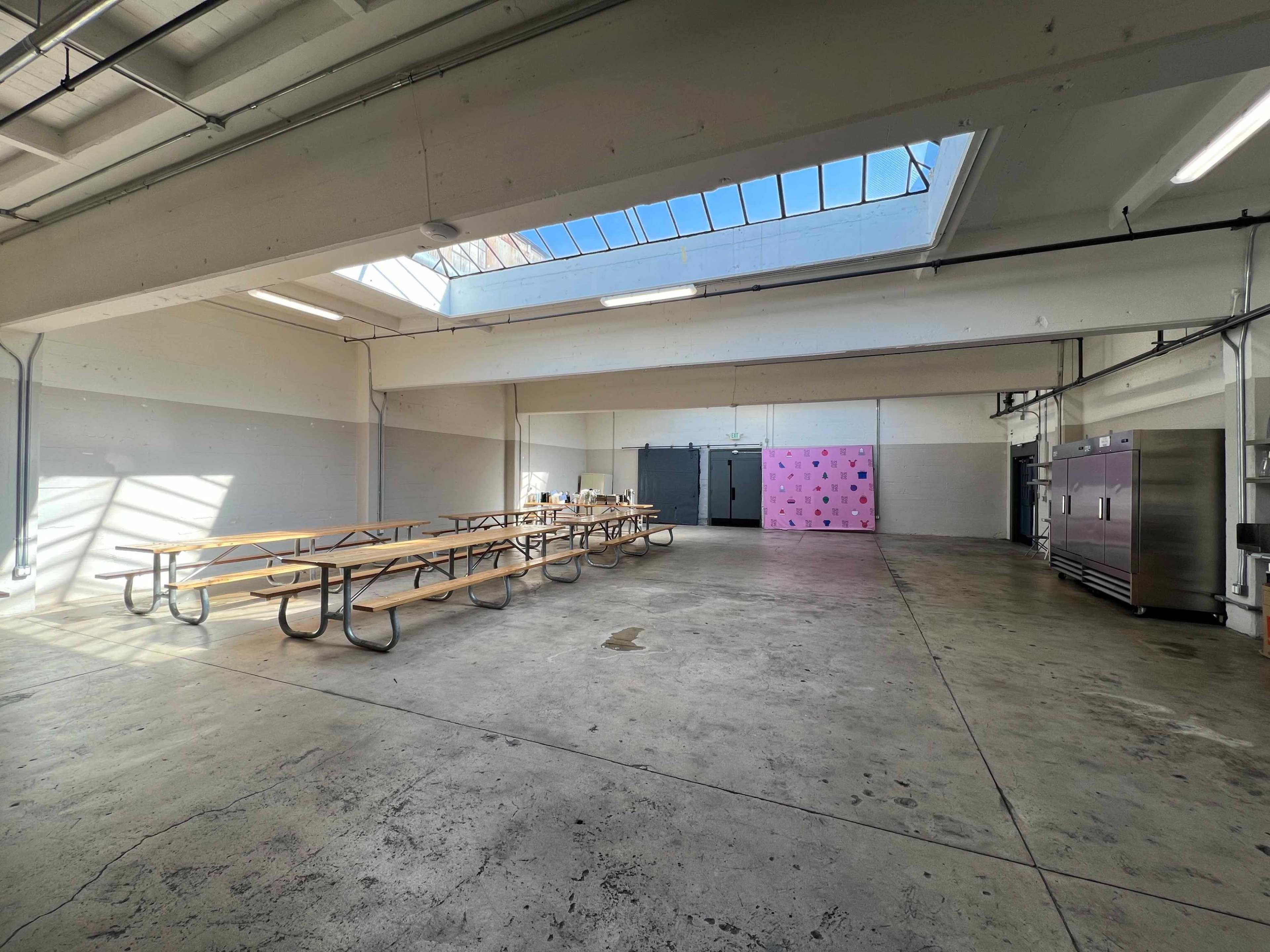 A spacious, empty room with rows of wooden picnic tables, a pink wall at one end, and large refrigerators along the side.