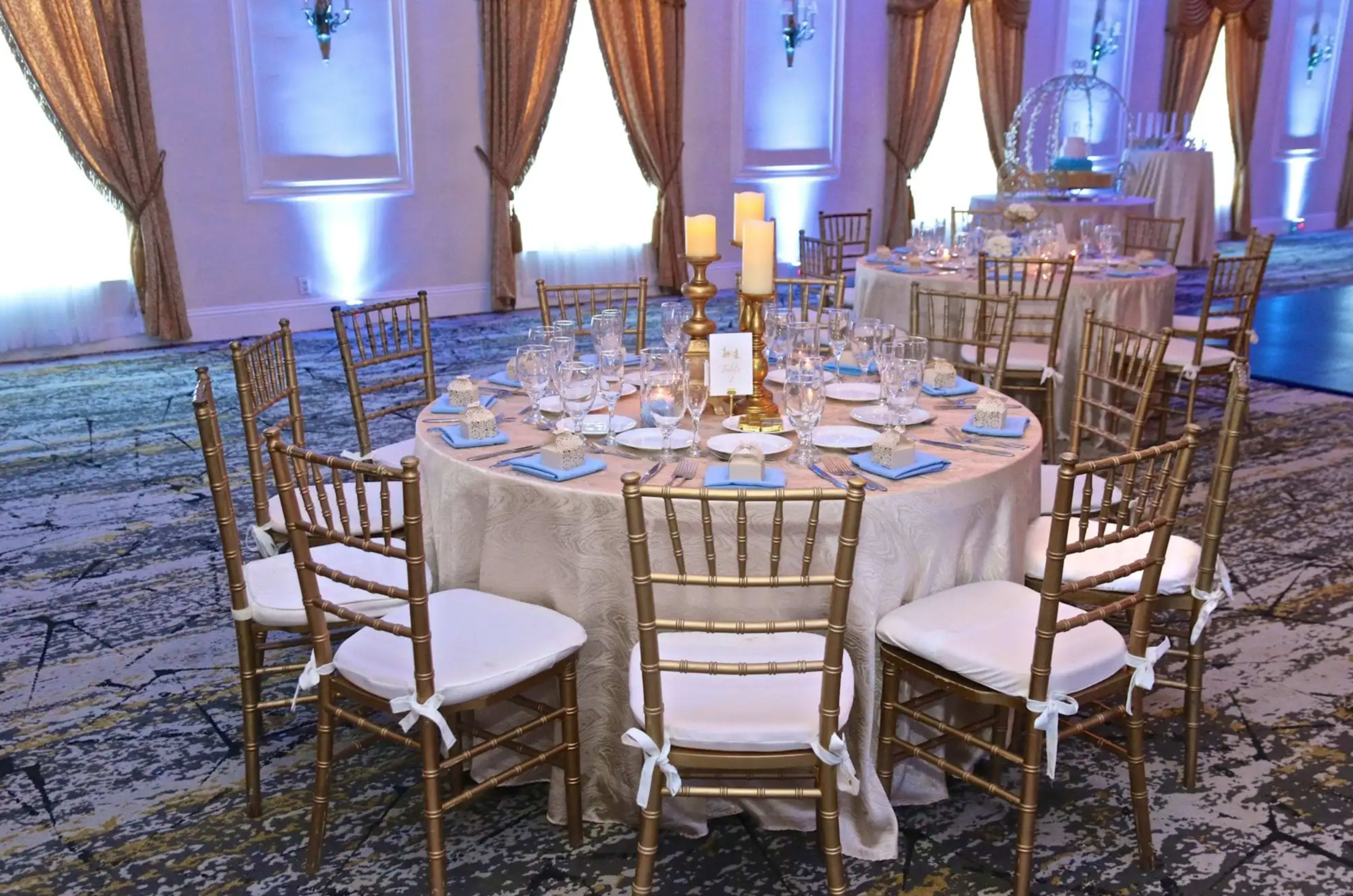 A round banquet table is elegantly set with glassware, plates, and a gold candle holder, surrounded by gold Chiavari chairs in a well-lit event space.