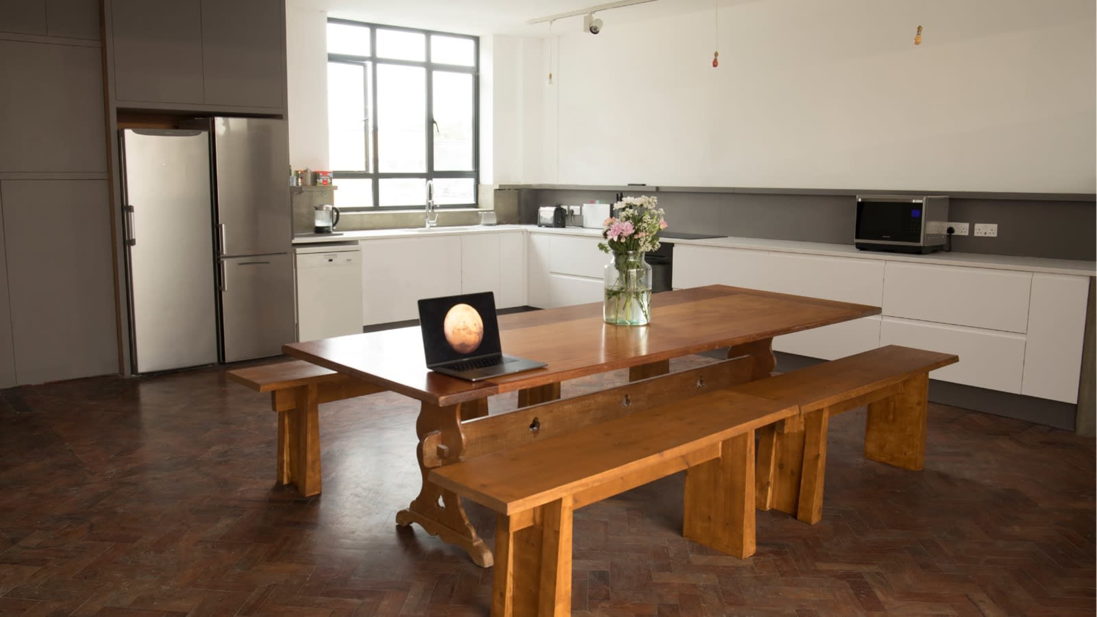 The image shows a modern kitchen with a large wooden table, benches, a laptop on the table, and kitchen appliances in the background.