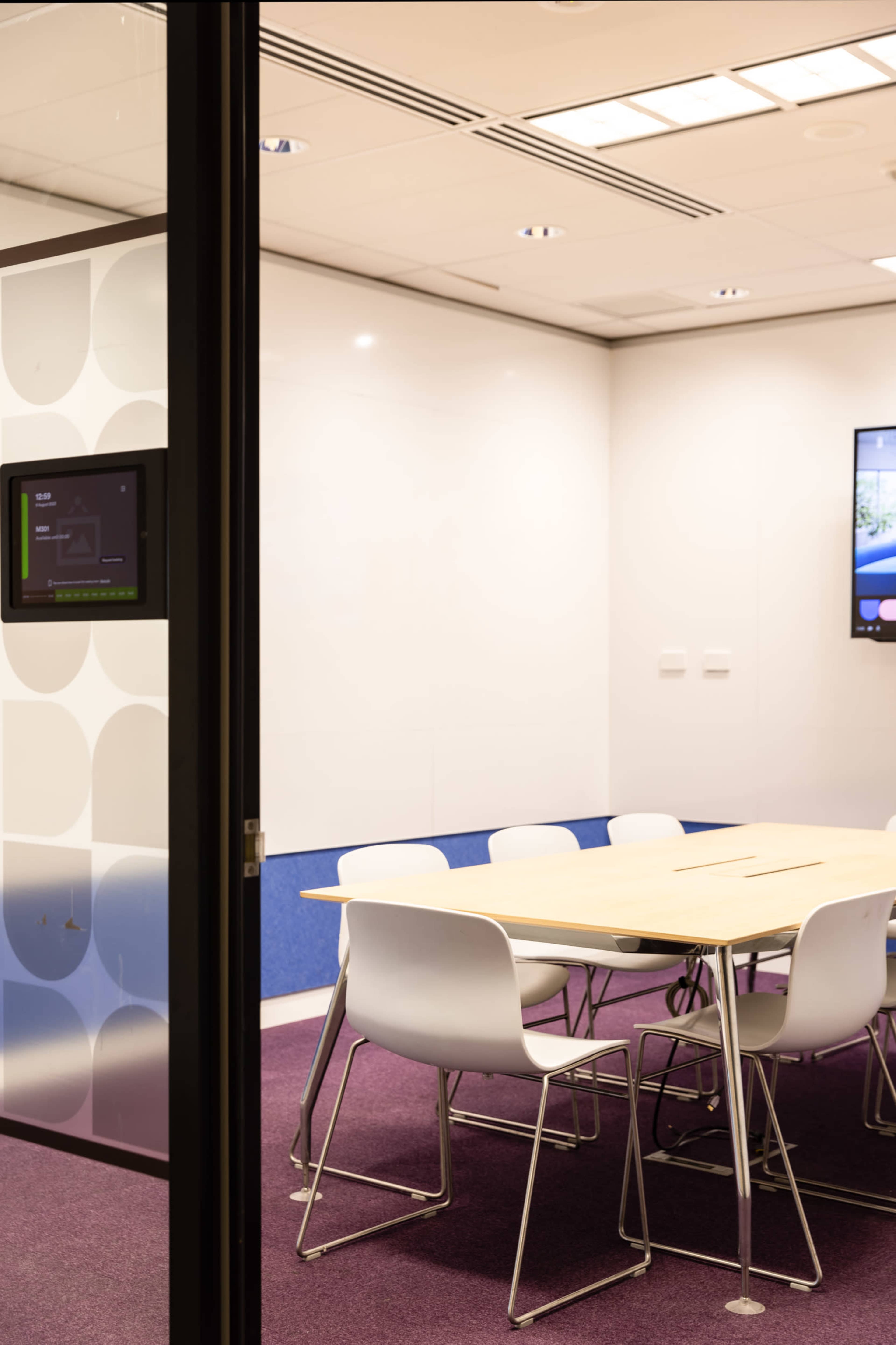 A modern conference room with a large wooden table surrounded by white chairs, featuring a digital display on one wall.