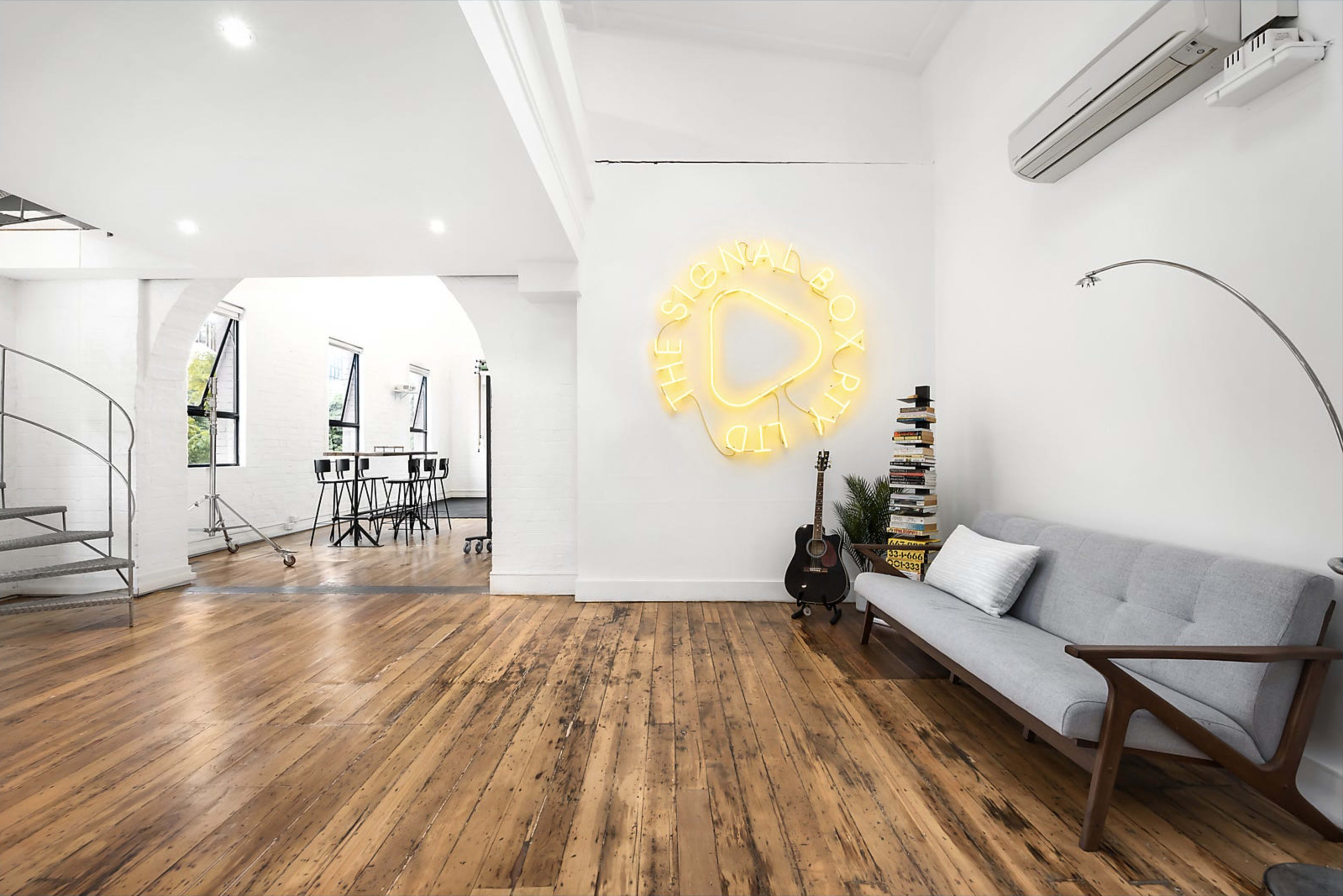 The image displays a spacious, well-lit room featuring a gray sofa, a neon sign on the wall, and a glimpse of a modern workspace in the background.