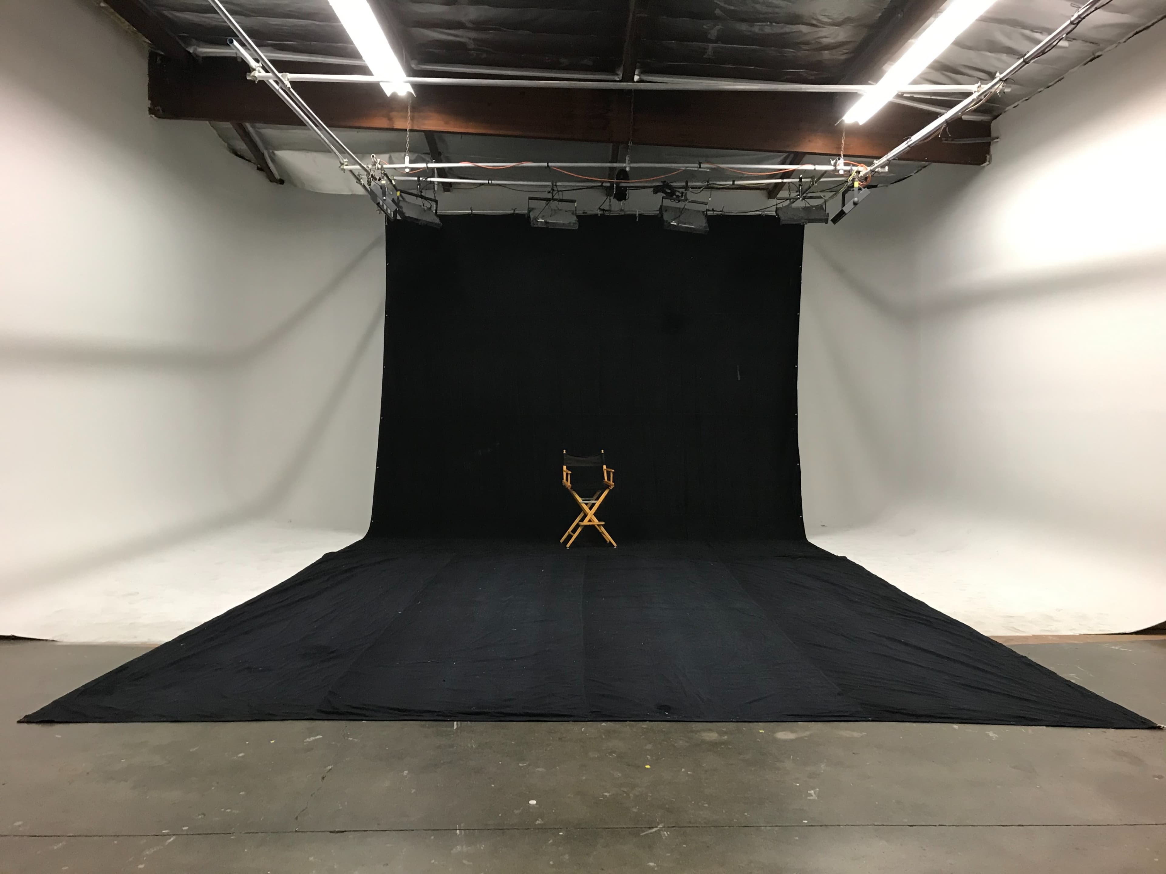 A wooden director's chair sits in the center of a photography studio with black backdrops and a concrete floor.