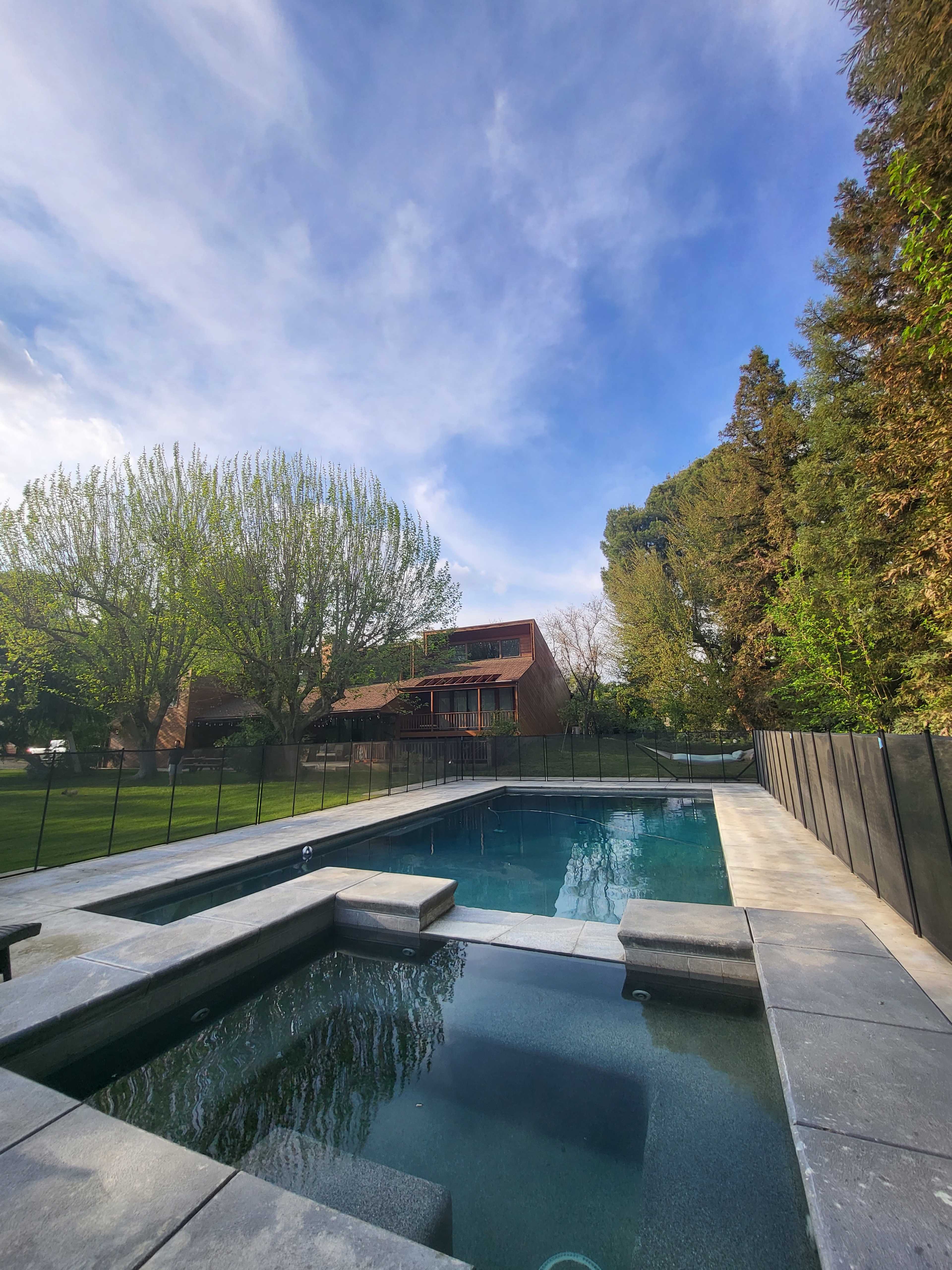 A rectangular pool with a connected spa, surrounded by a black fence, grassy areas, and trees, under a clear sky.