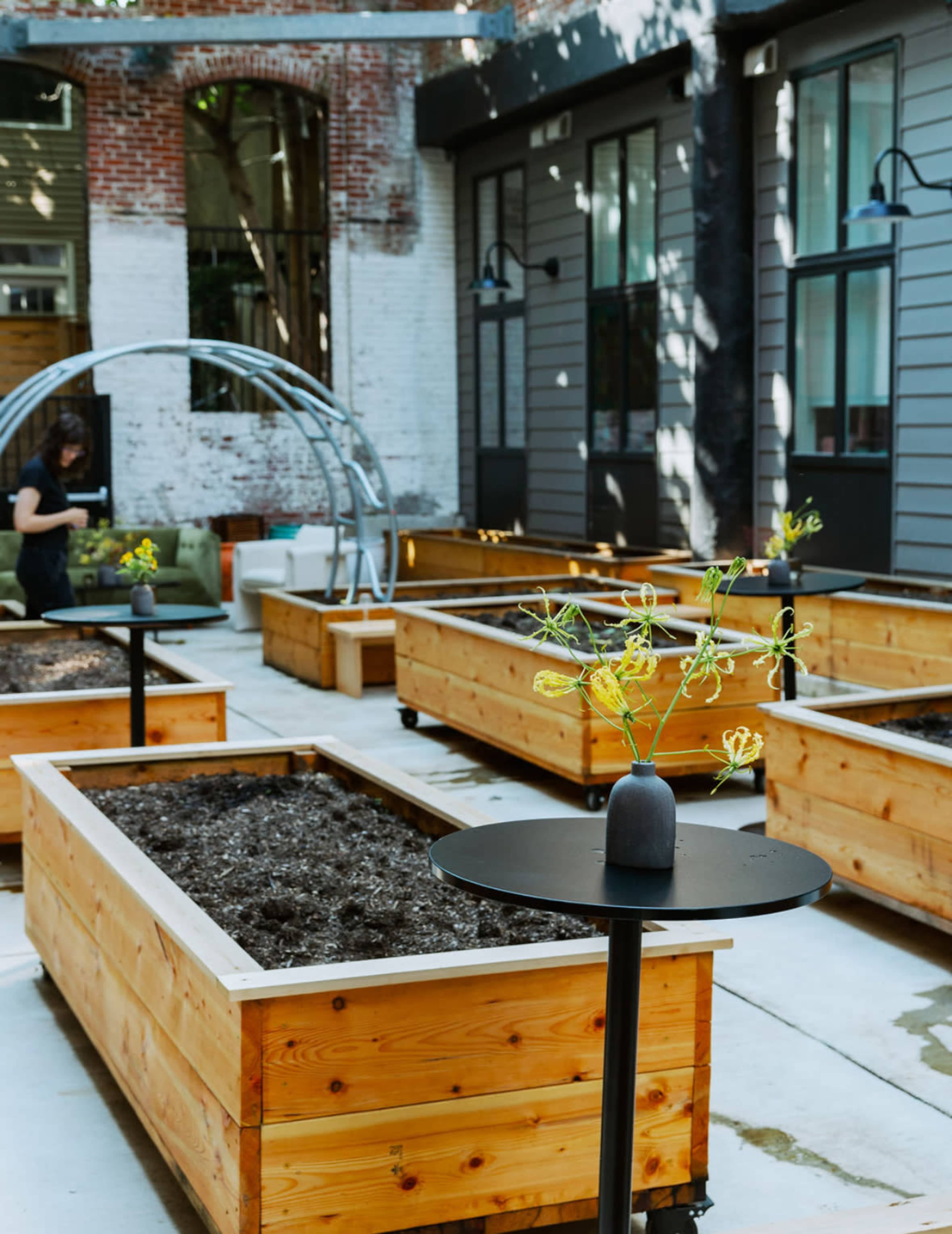 The image shows a courtyard with wooden raised garden beds, a table with a vase of flowers, and a person tending to a nearby area.
