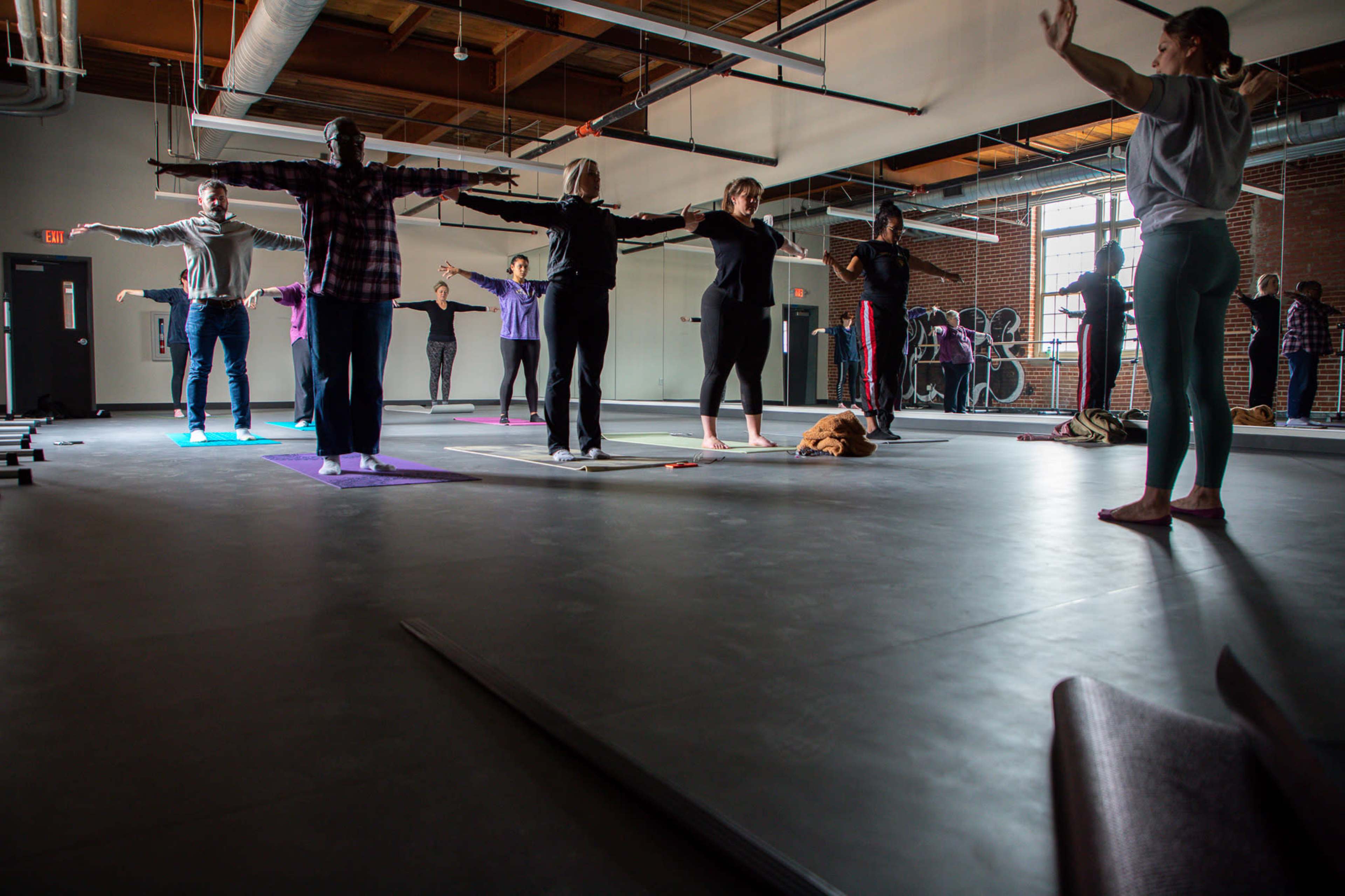 A group of people is practicing yoga in a spacious studio with large windows and wooden beams overhead.
