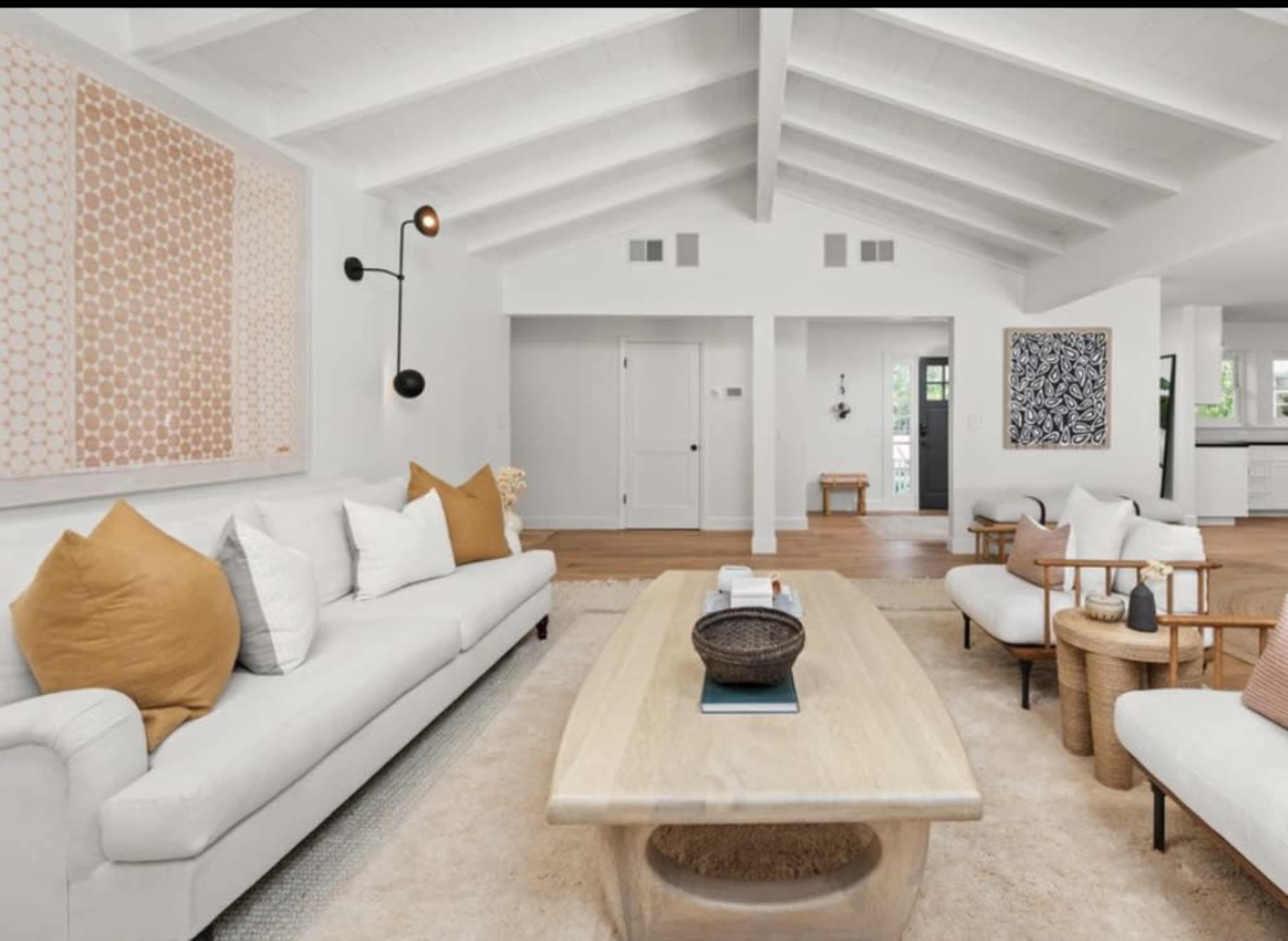 The living room features a white sofa with brown and gold accent pillows, a wooden coffee table, and a light-colored area rug under a beamed ceiling.