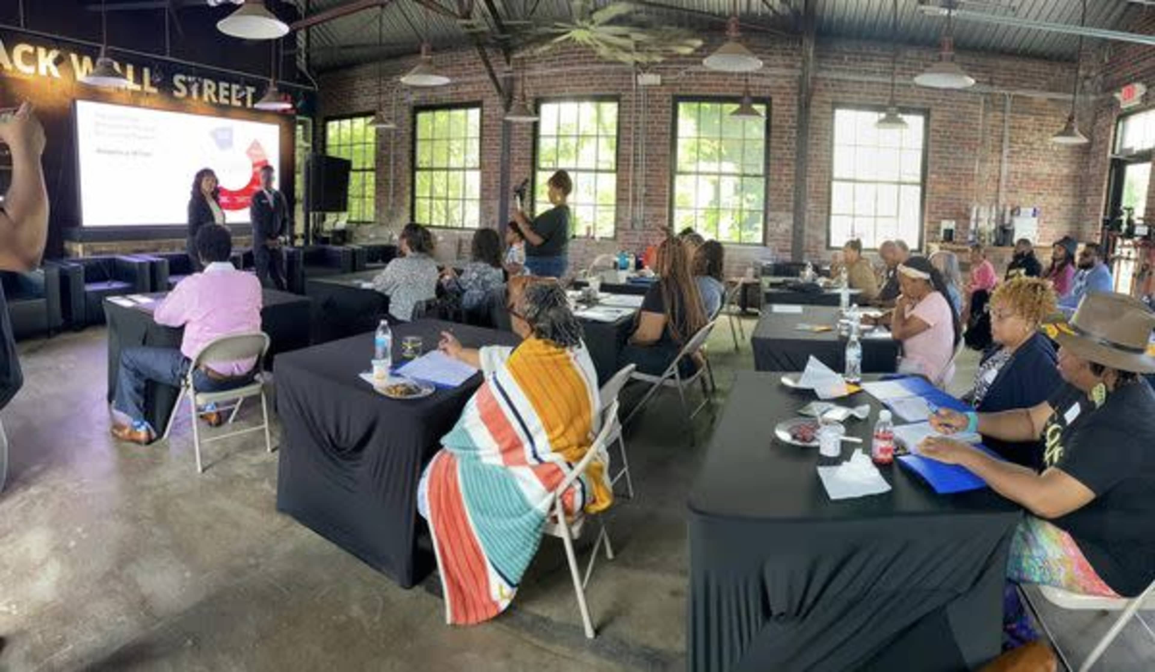 A group of people are seated at tables in a large room with exposed brick walls, attending a presentation in front of a screen.