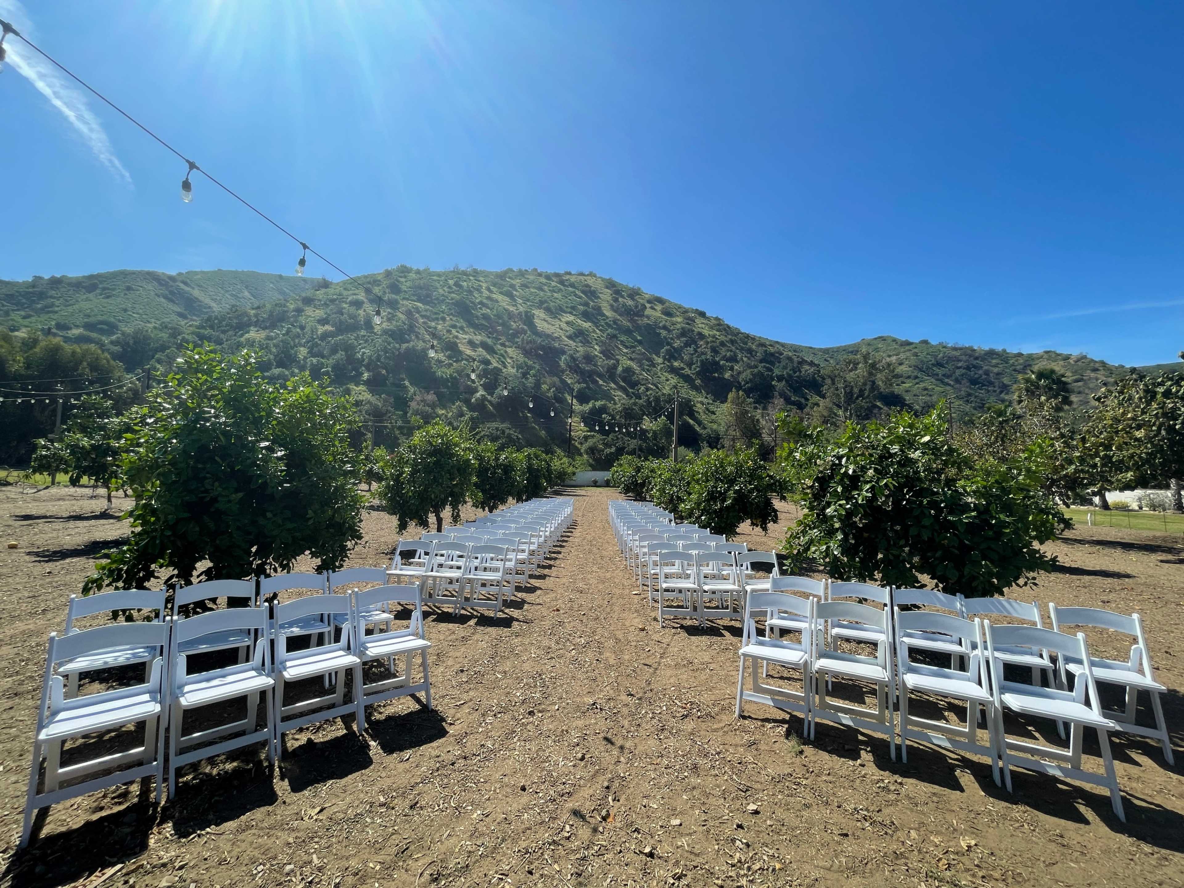 A wedding ceremony is set up outdoors with white chairs arranged in rows on dirt, flanked by trees, against a backdrop of a green hillside.