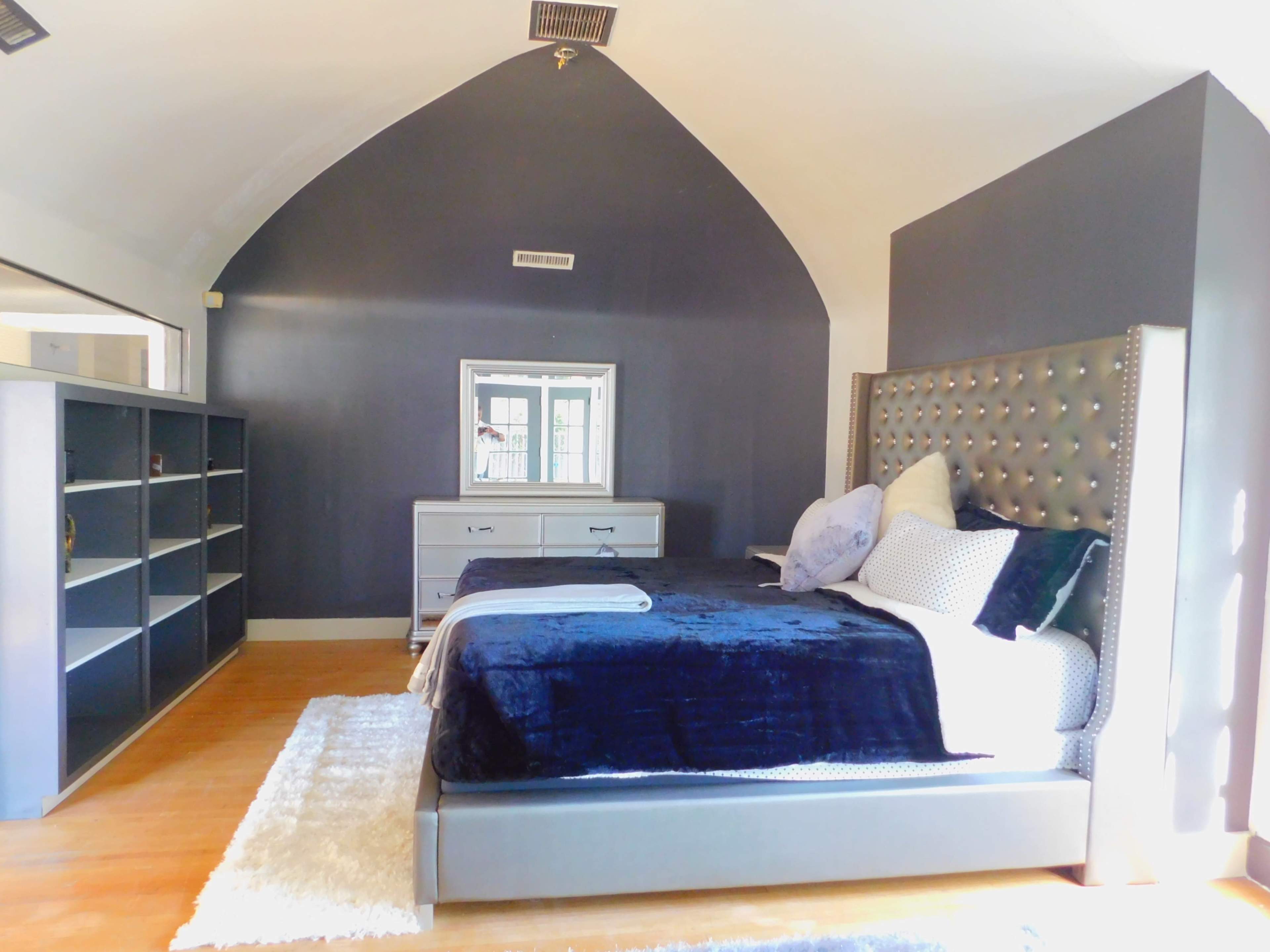 The bedroom features a large bed with a tufted headboard, a mirrored dresser, and a shelving unit against a dark wall.