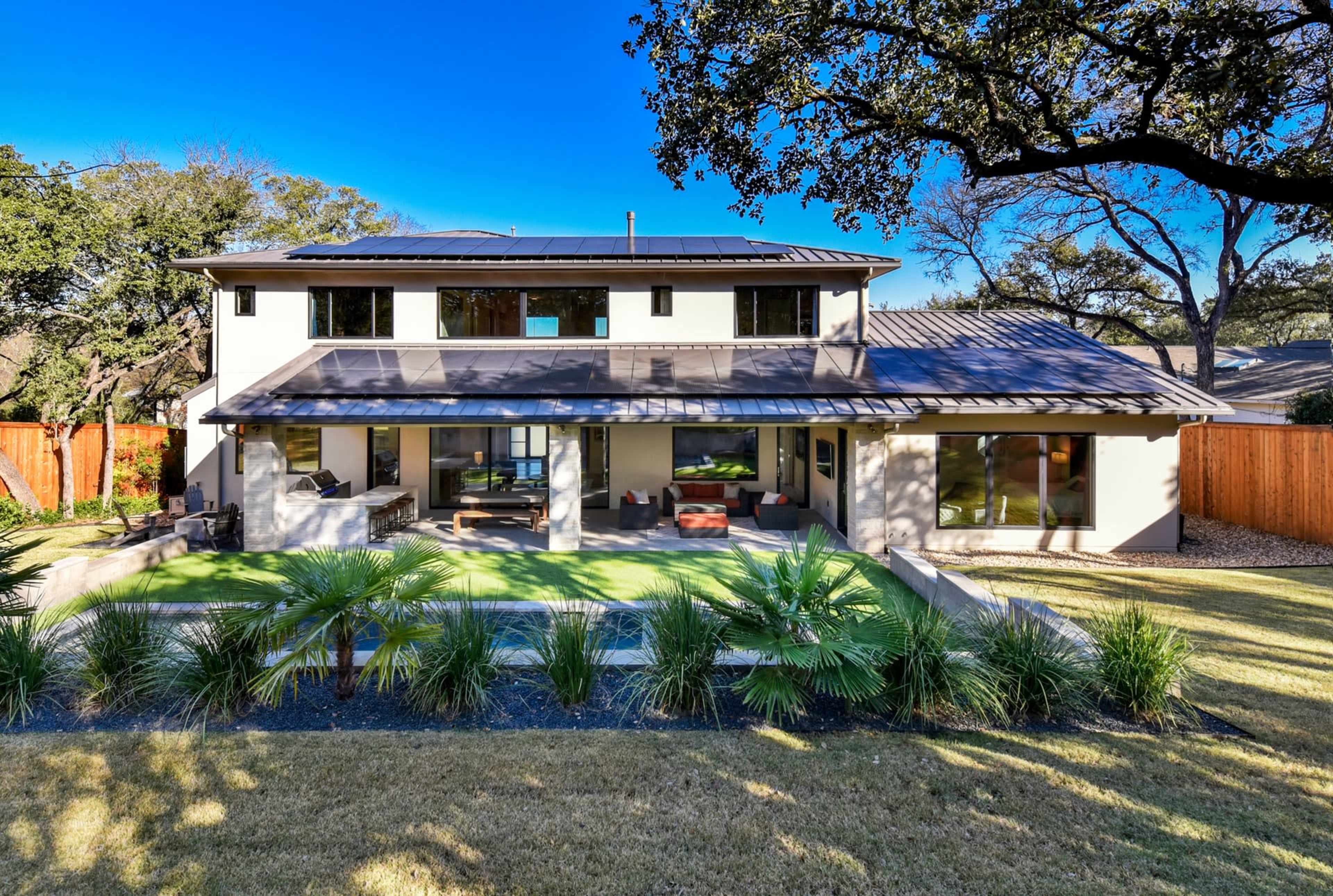 The image shows a modern two-story house with a metal roof, large windows, and a landscaped yard featuring palm plants.