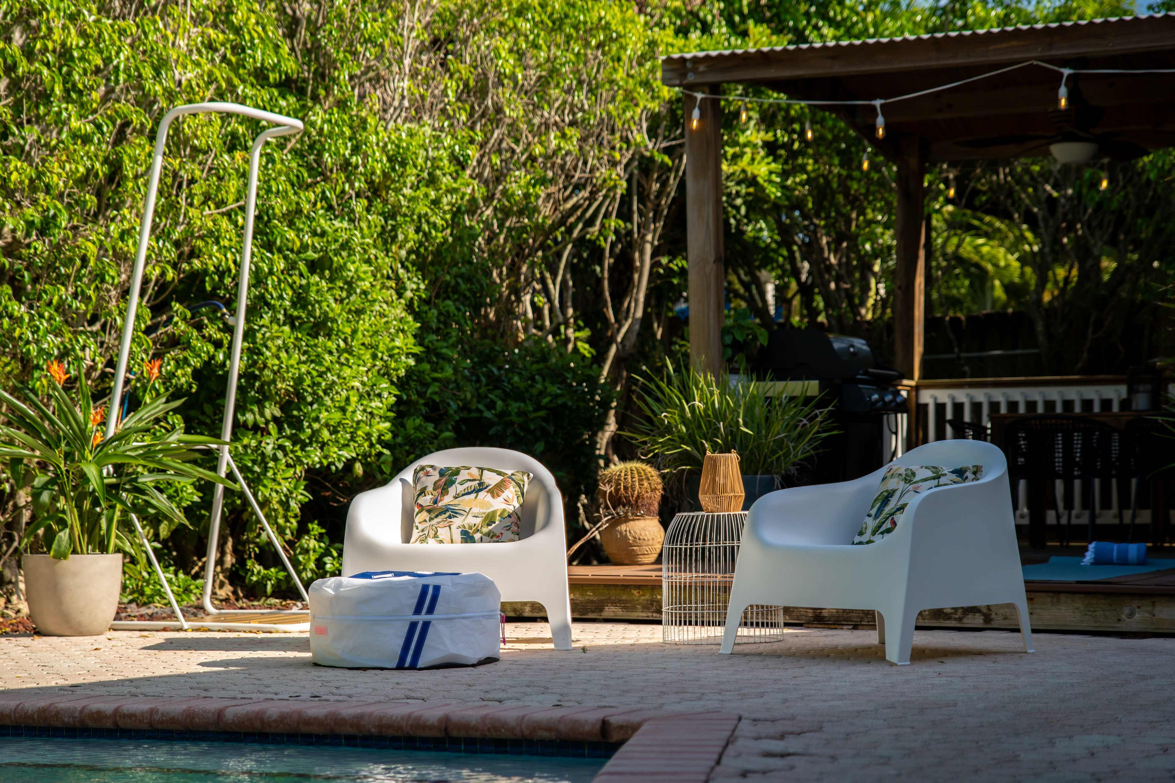 Two white outdoor chairs with patterned cushions are positioned beside a pool near a shaded patio area surrounded by greenery.