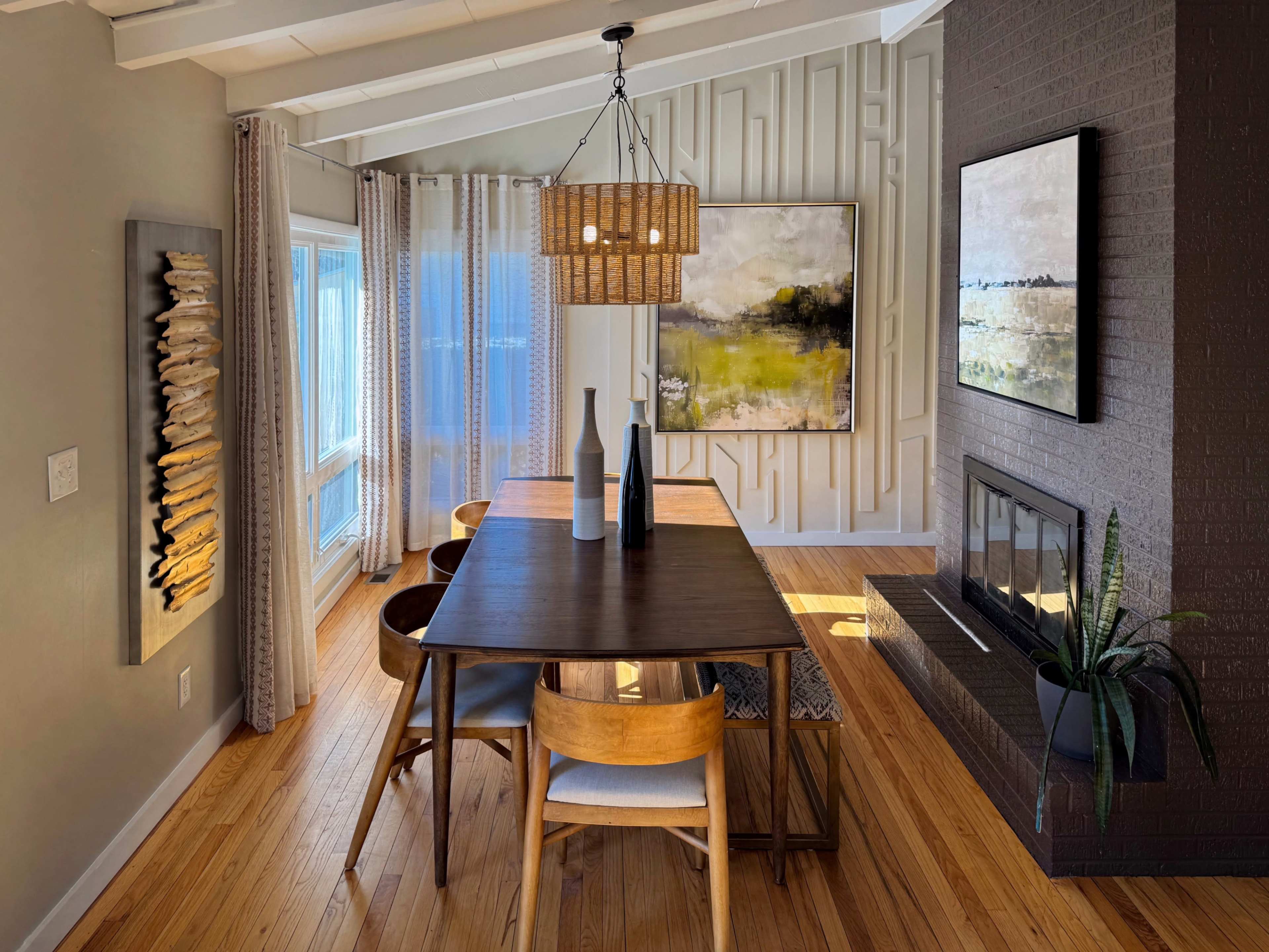 A modern dining area features a wooden table with chairs, a large painting on the wall, and a cozy fireplace, all illuminated by natural light from windows.