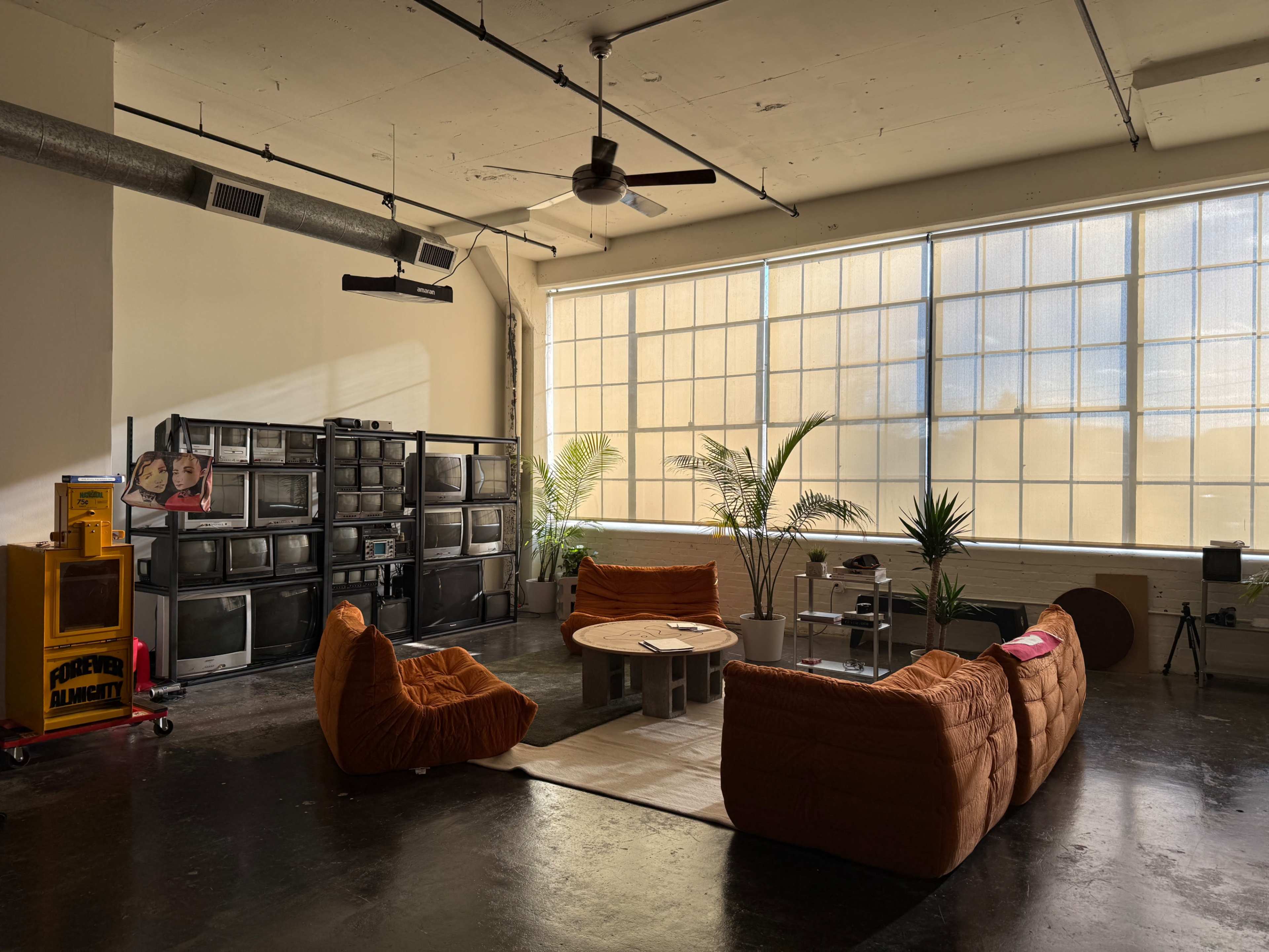 The scene showcases a spacious, well-lit room with large windows, featuring a collection of vintage televisions, a round coffee table, and cozy orange lounge furniture surrounded by potted plants.