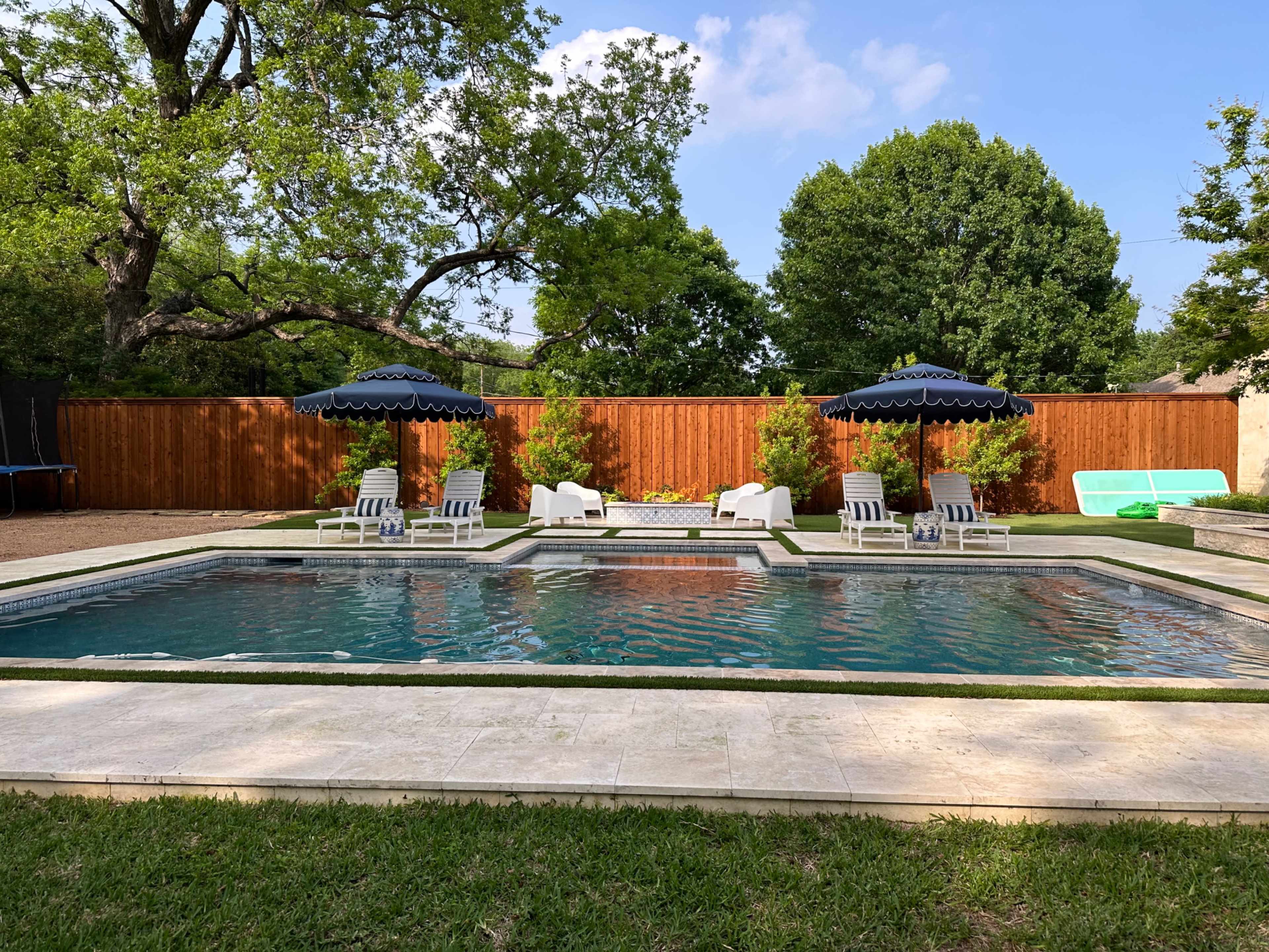 A backyard pool area surrounded by green grass, featuring lounge chairs, two umbrellas, and a wooden fence in the background.
