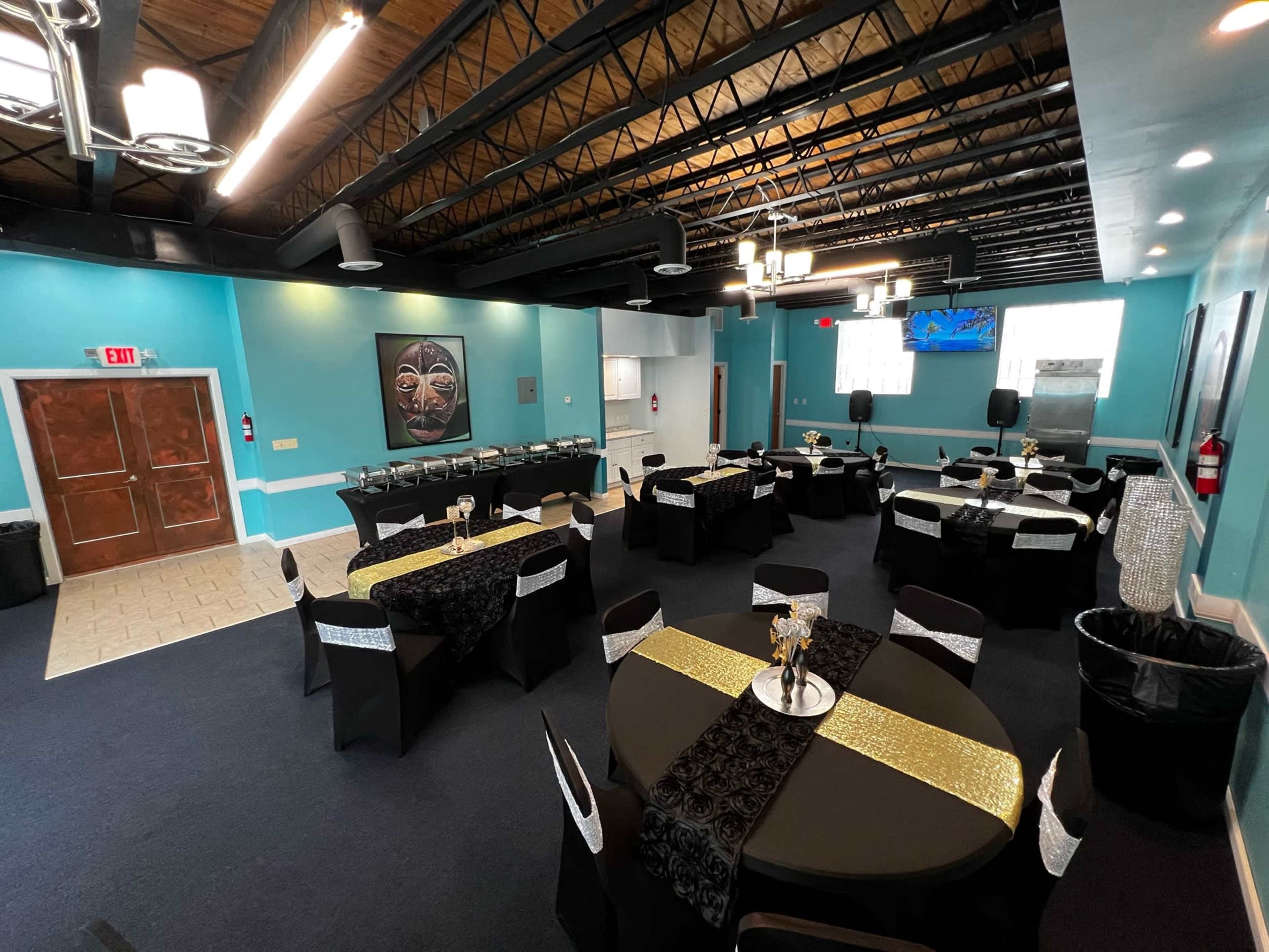 A spacious event room with several round tables covered in black and gold tablecloths, set up for a gathering, and adorned with decorative centerpieces.