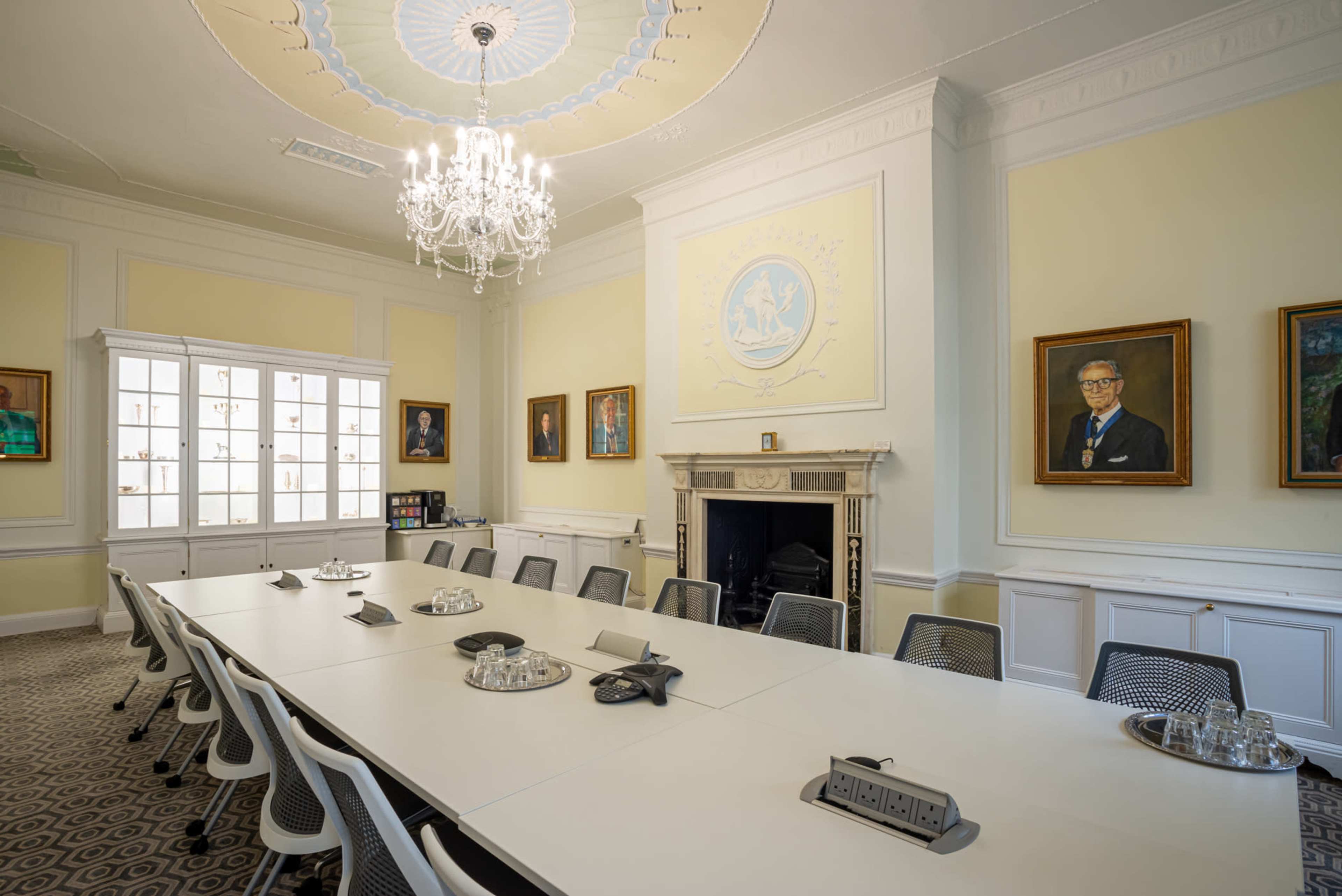 A formal conference room with a long white table, black and white chairs, a chandelier, and framed portraits on the walls.