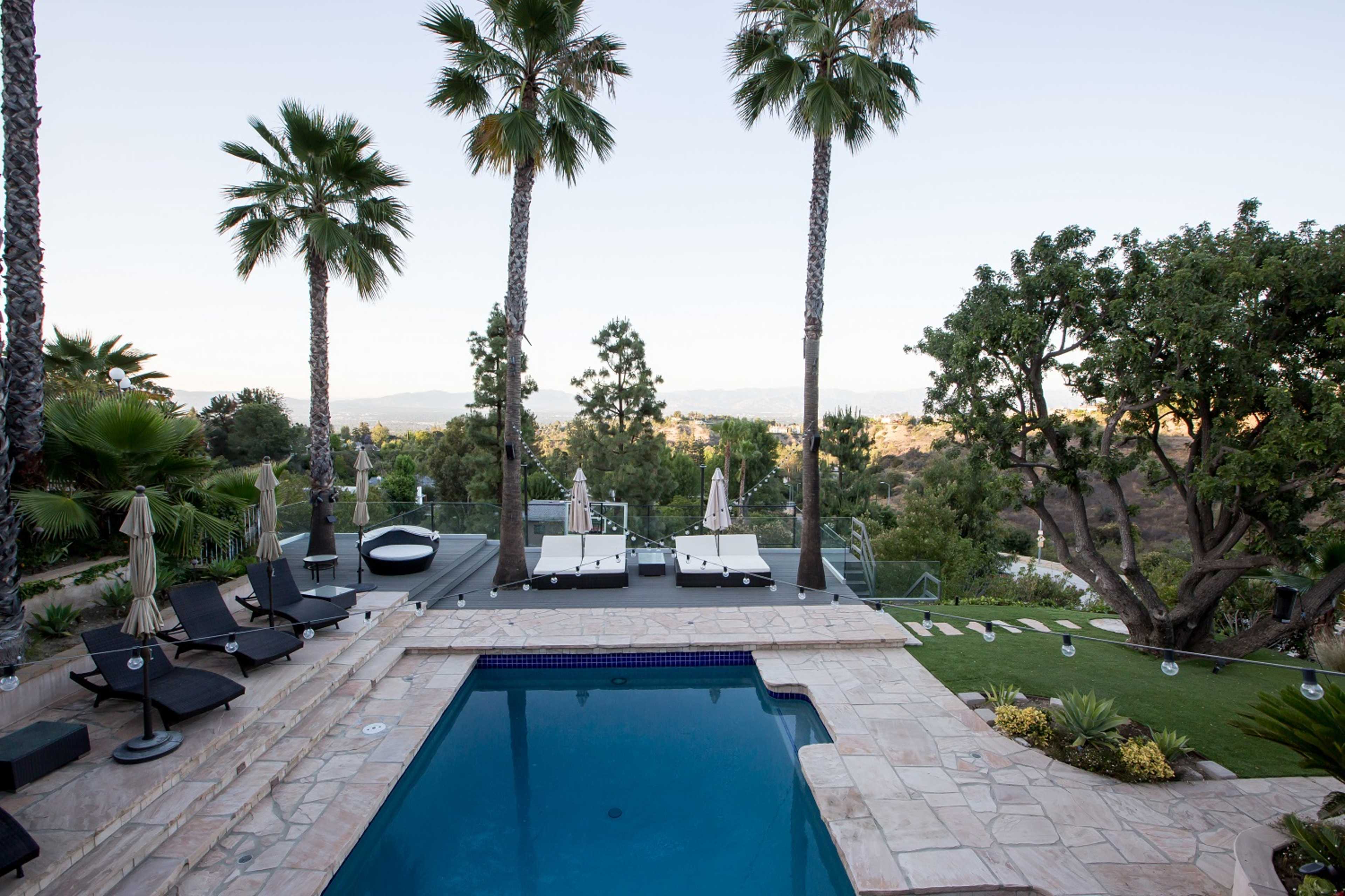 A backyard pool area surrounded by palm trees, lounge chairs, and a view of hills in the background.
