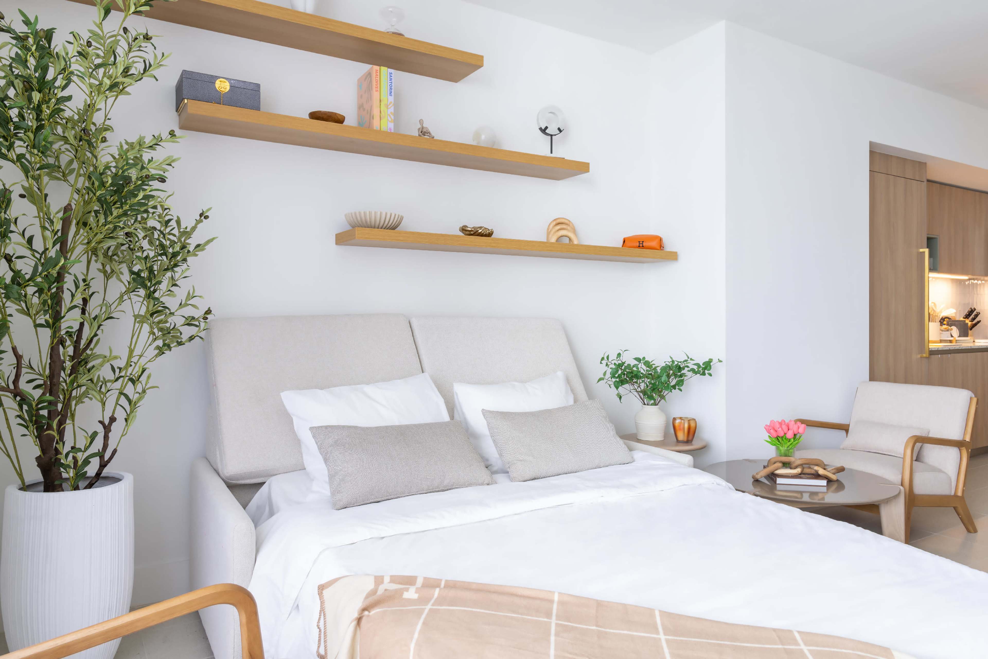 The image shows a modern bedroom featuring a light-colored bed with two decorative pillows, minimalistic shelves above, and a small seating area with a plant and a vase of flowers.