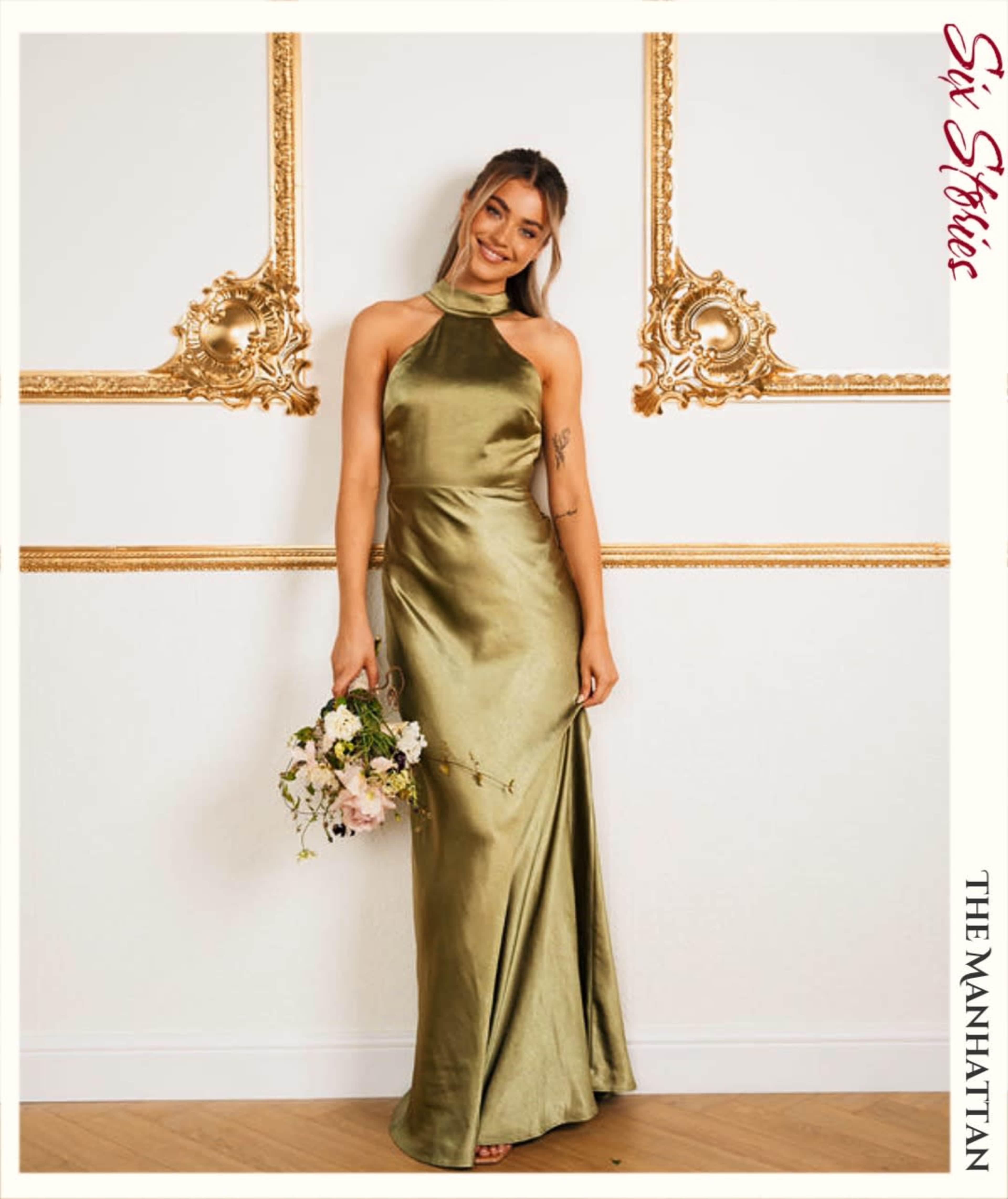 A woman wearing a satin olive green dress holds a bouquet of flowers in front of a decorative gold-accented wall.