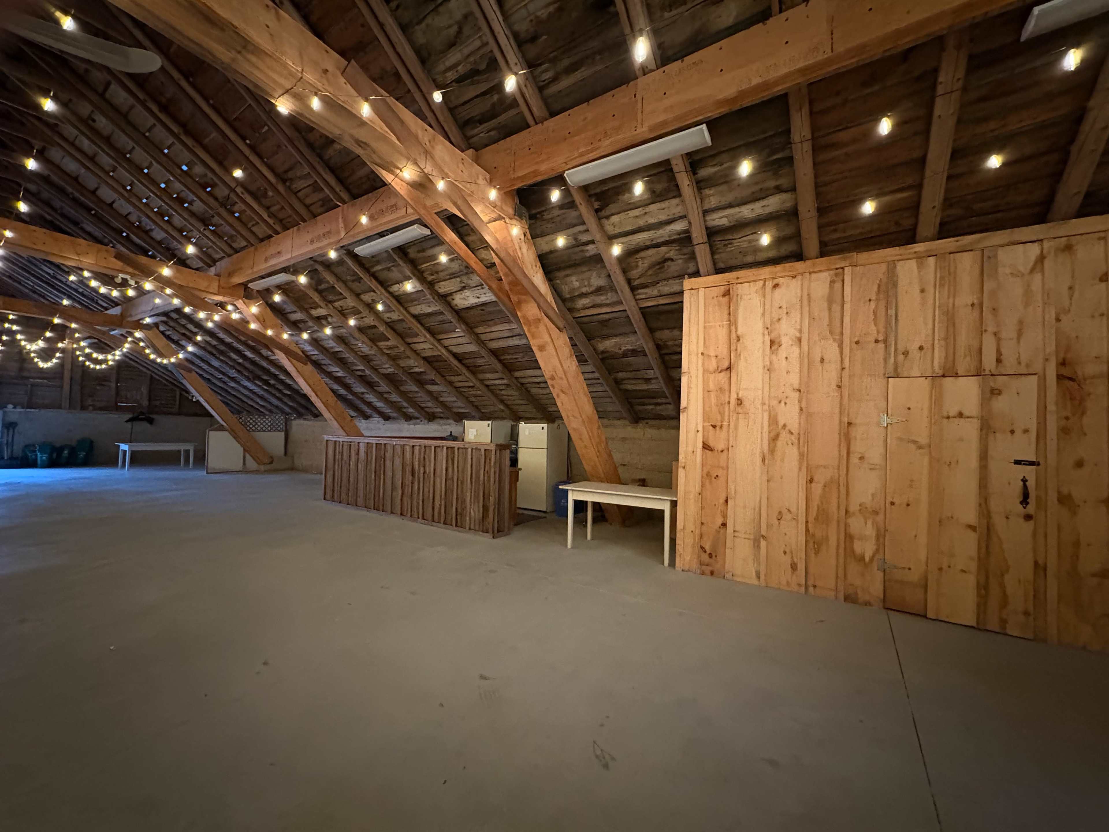 The image shows a spacious attic with wooden beams and string lights, featuring a wooden bar area and a storage closet in one corner.