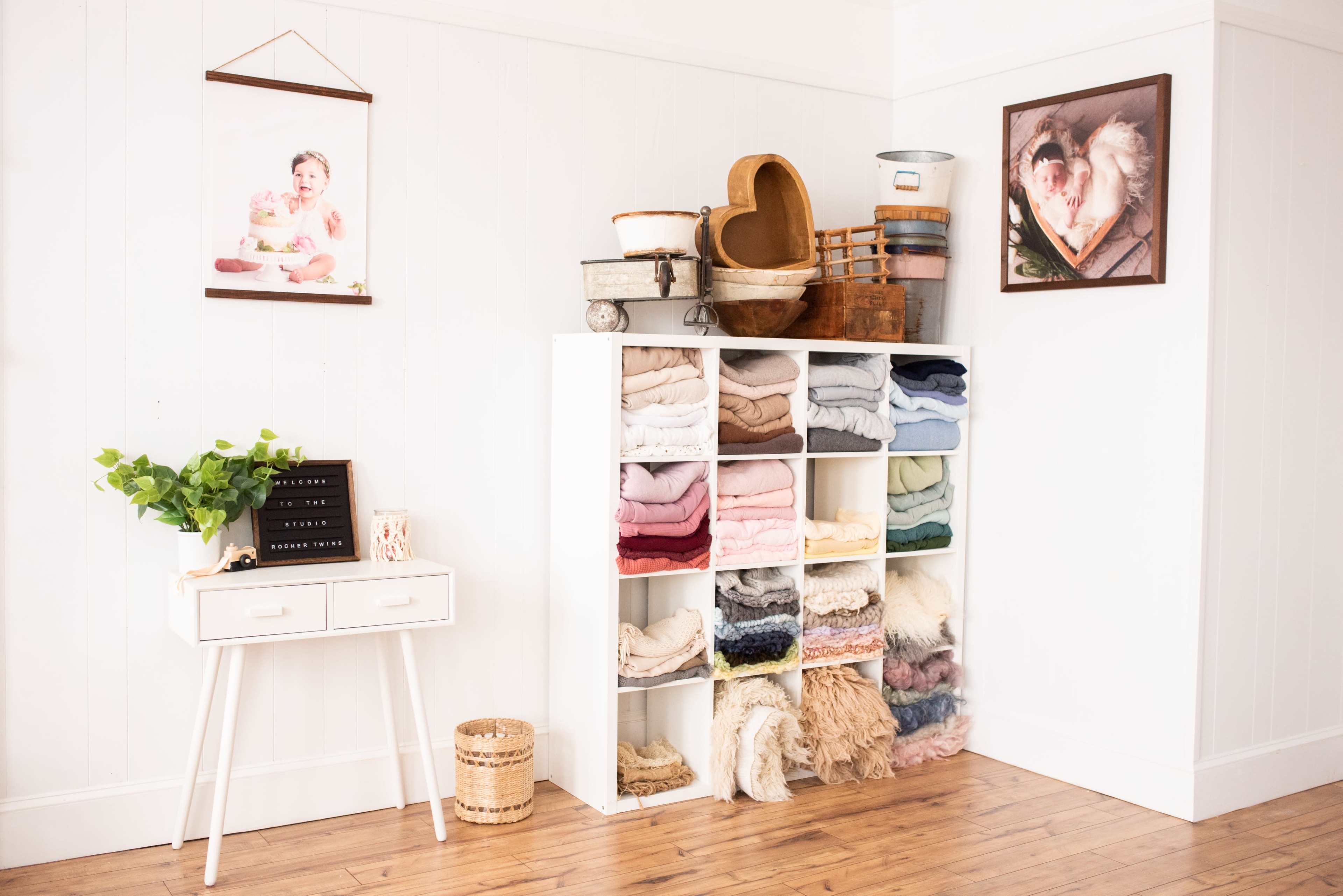 A corner of a room features a white shelf filled with neatly folded blankets, a small table with decorative items, and various woven baskets displayed above.