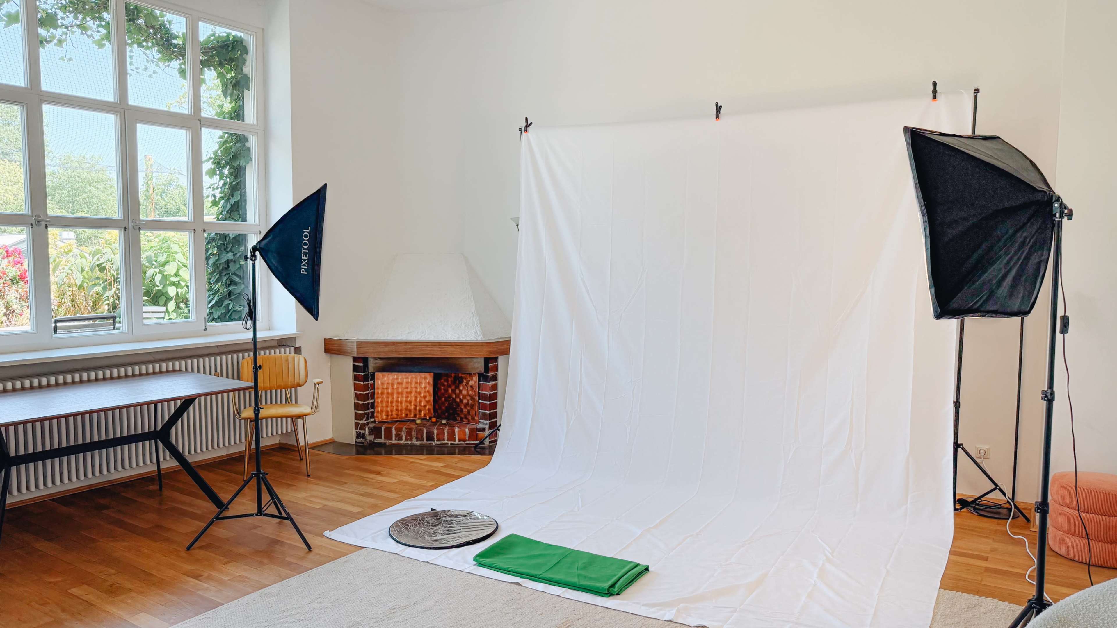 A clean studio space features a white backdrop, lighting equipment, and a wooden table with a chair.