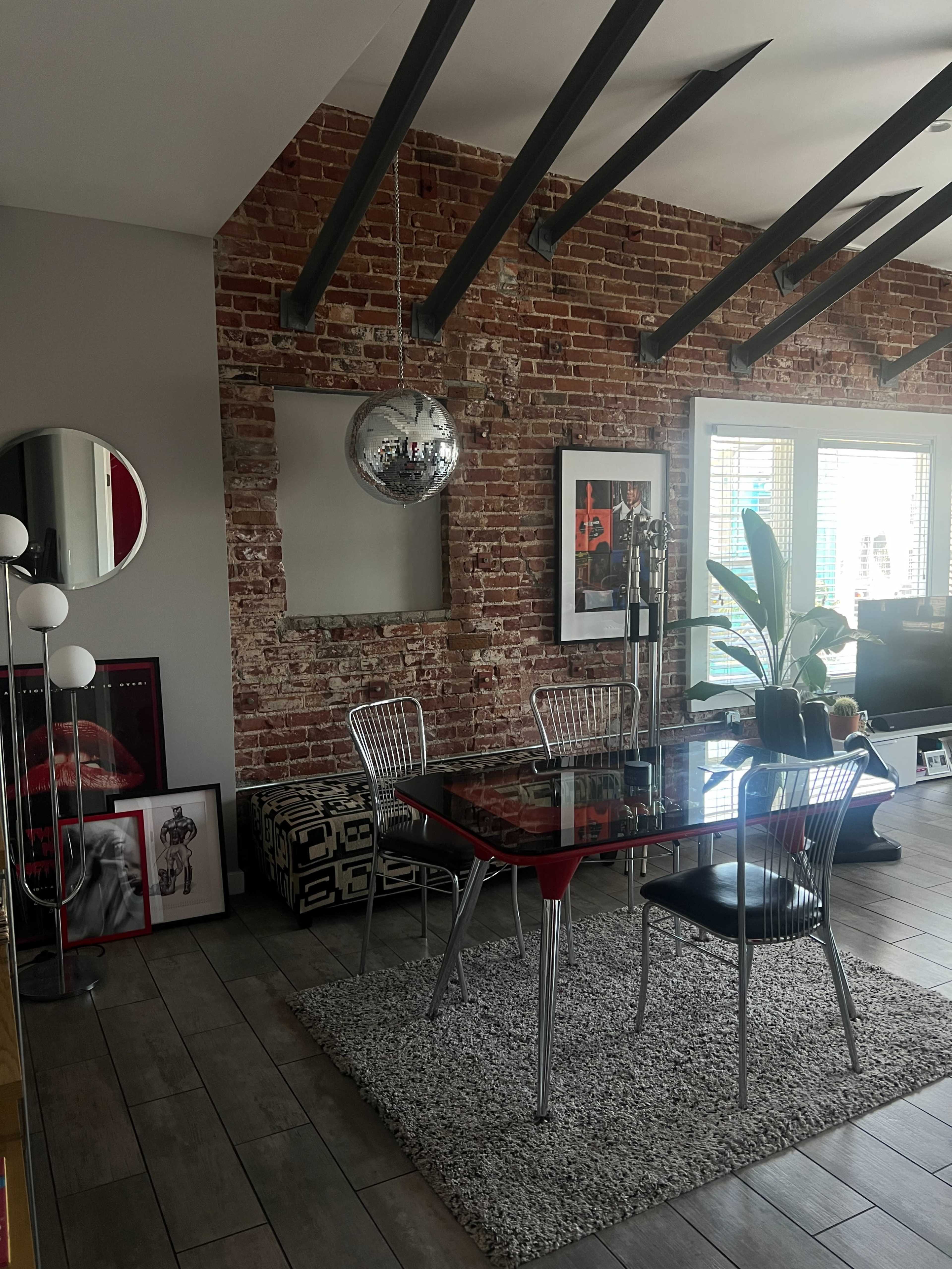 The image shows a modern dining area featuring a glass table with metal chairs, a disco ball overhead, and a brick wall with framed artworks and potted plants.