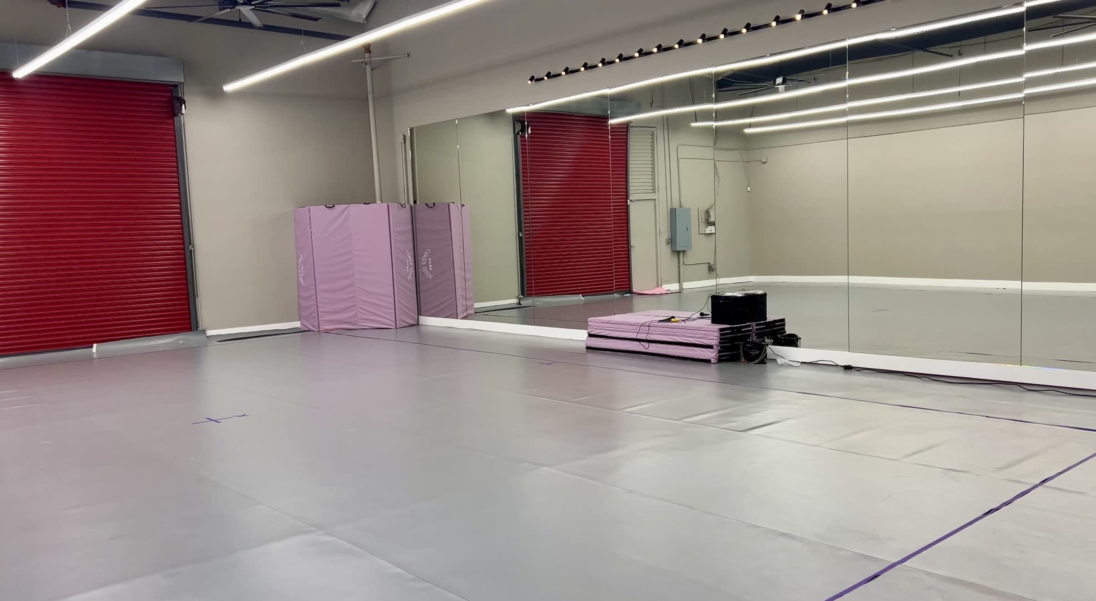 The image shows an empty dance studio with gray flooring, red rolling doors, and a large mirror along one wall.