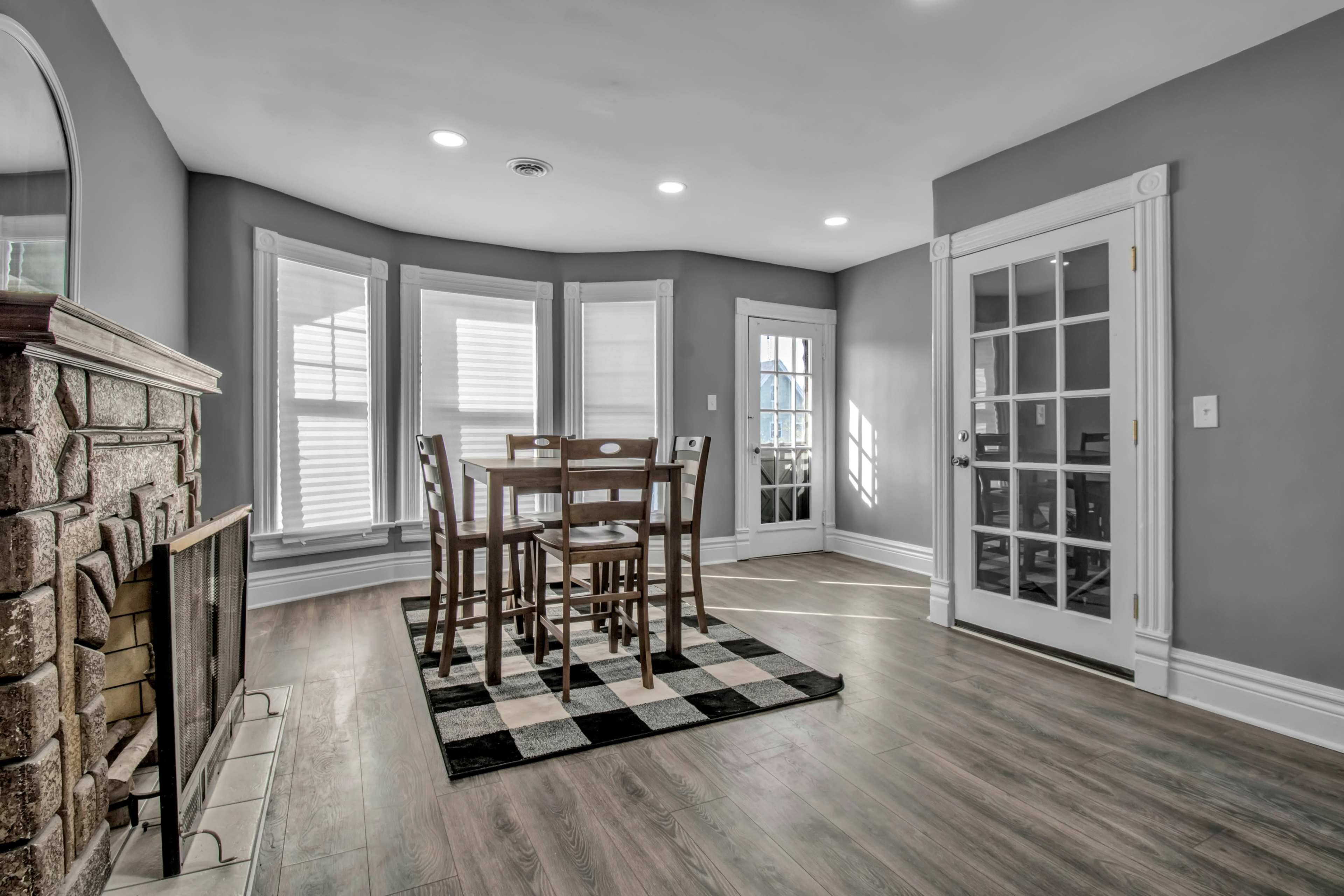 A well-lit dining area with a wooden table and chairs, set on a checkered rug next to a stone fireplace.