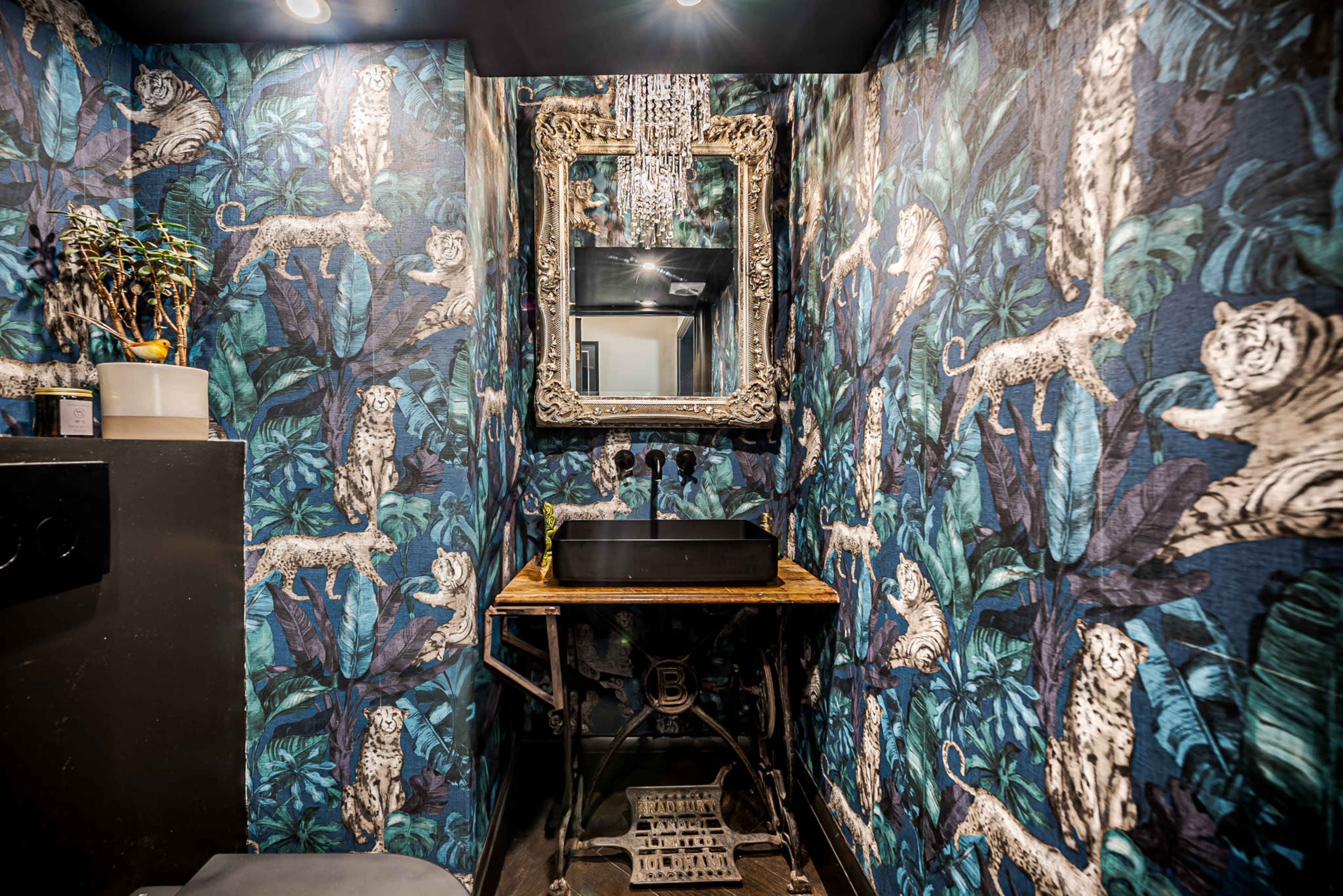 A small bathroom features a black sink atop a wooden table, surrounded by vibrant animal-patterned wallpaper and a decorative mirror.