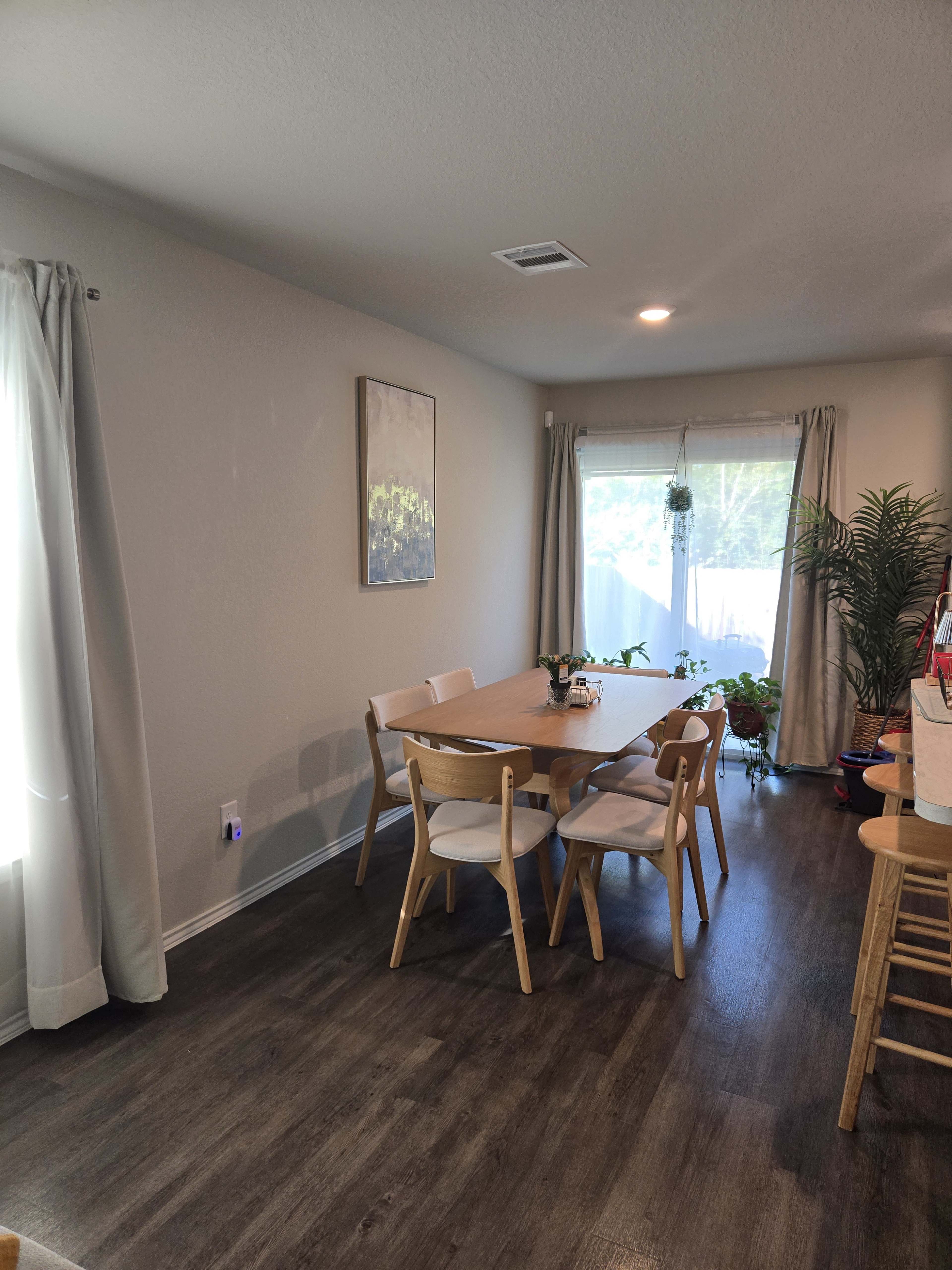 A dining area features a wooden table surrounded by white chairs, with a window and potted plants in the background.