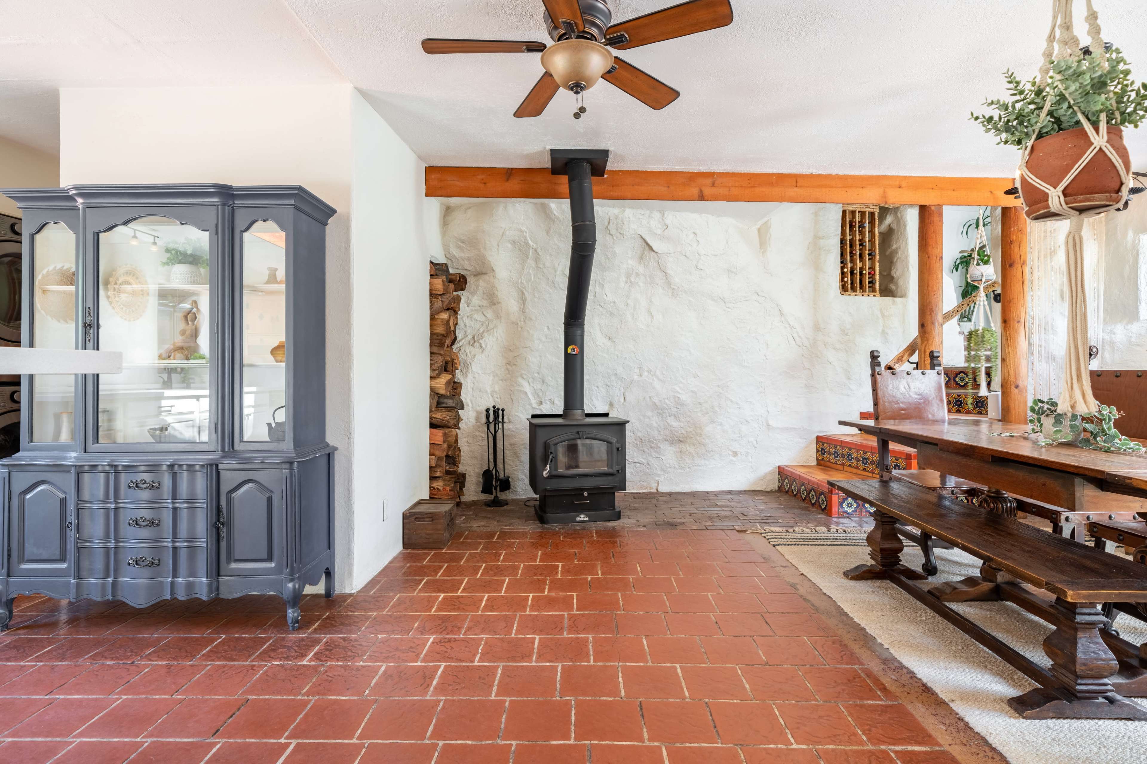 The image shows a cozy living area featuring a wood-burning stove, a wooden dining table, and a gray china cabinet against a white stone wall.
