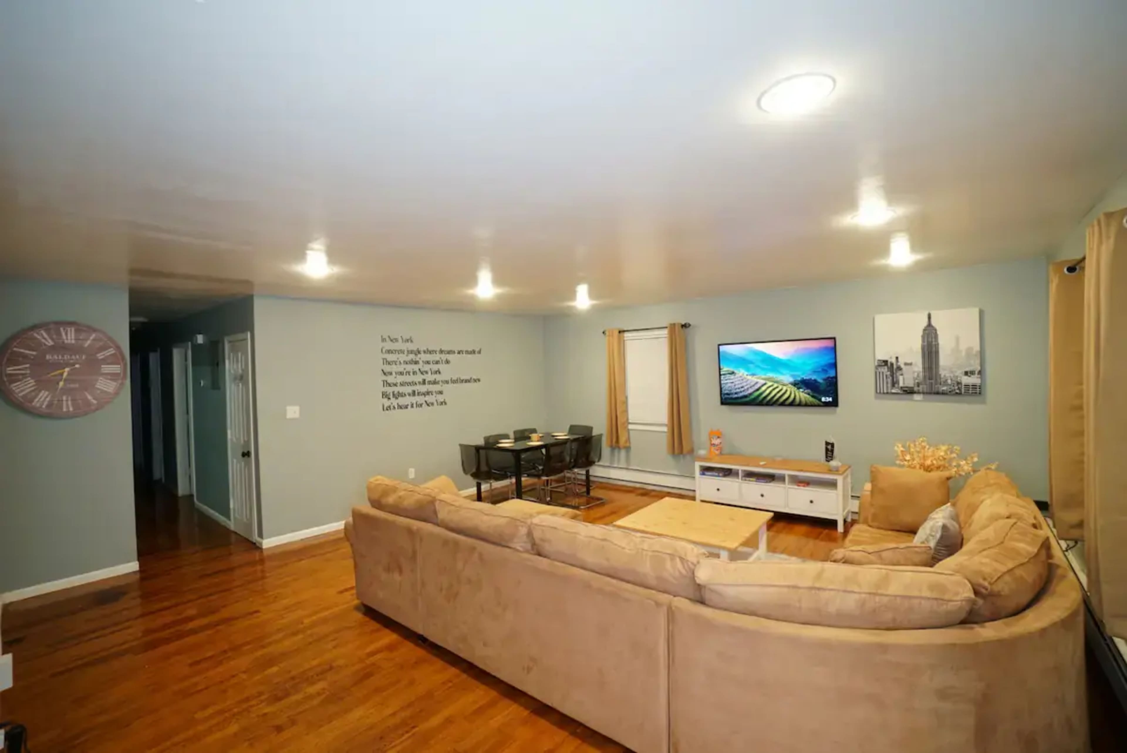 A spacious living room with a large beige sectional sofa, a wall-mounted television, and a dining table set against a blue wall with decorative elements.