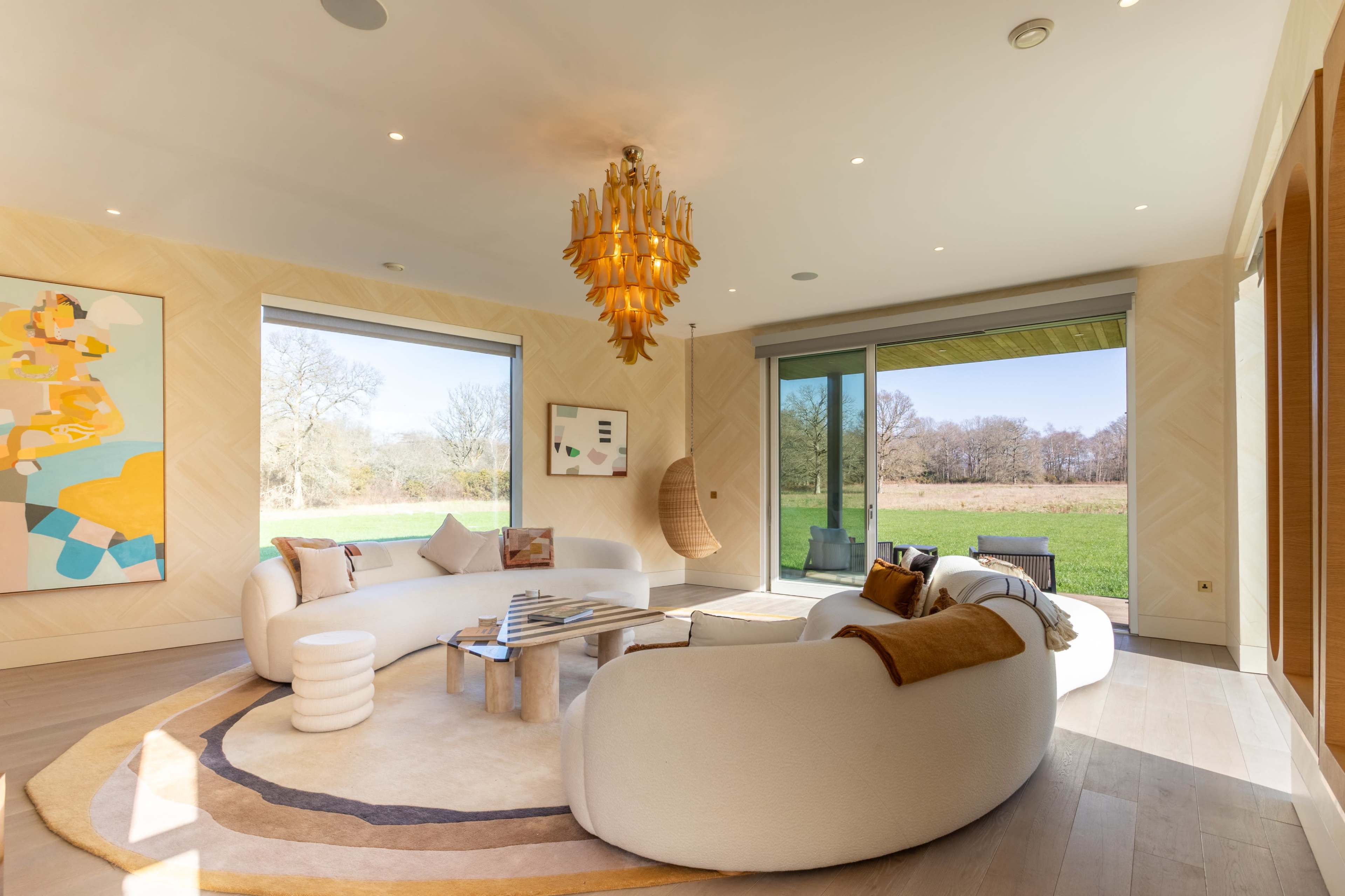 The image shows a modern living room with a large circular sofa, a coffee table, abstract art on the walls, and floor-to-ceiling windows overlooking a green landscape.