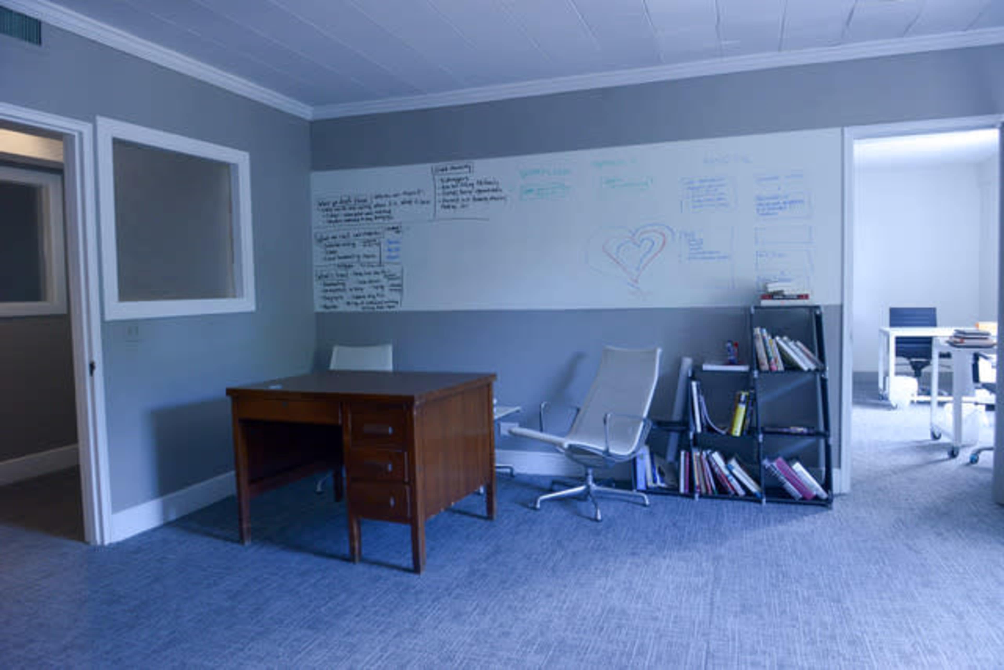 A minimally furnished office with a wooden desk, an ergonomic chair, and a whiteboard covered in notes on the wall.