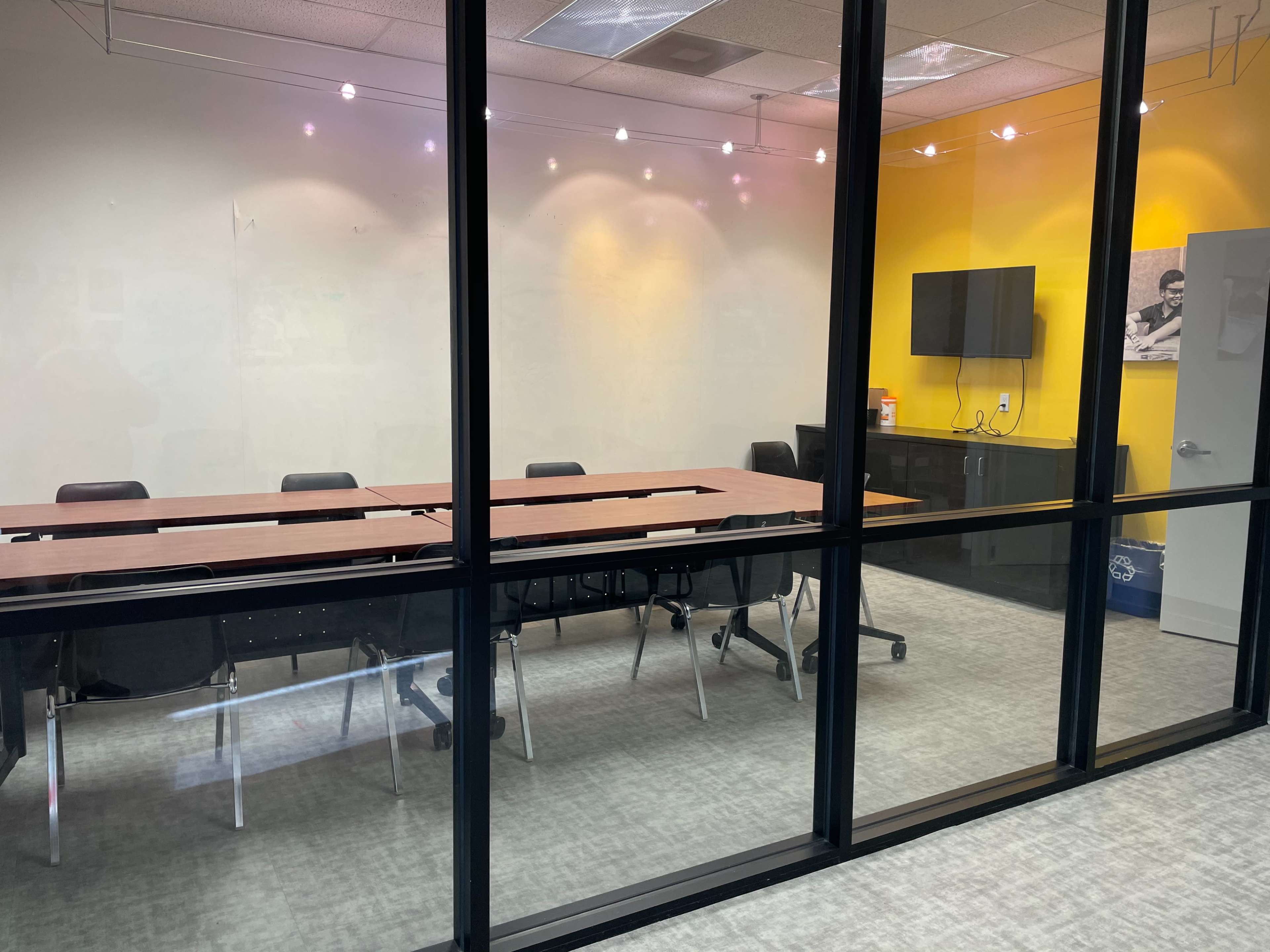 A conference room with a long wooden table and black chairs, surrounded by glass walls and a yellow accent wall.