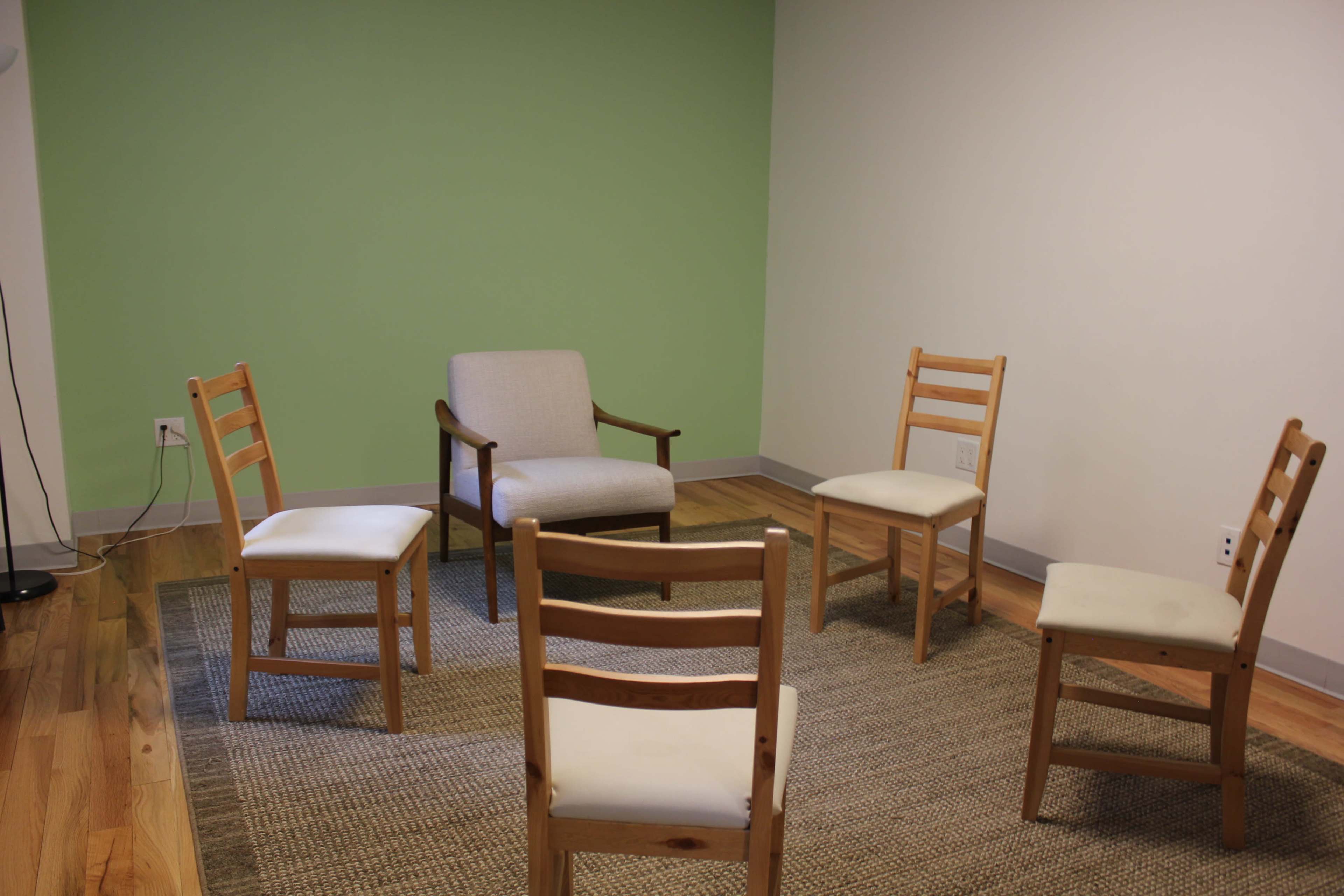 A small room with a green accent wall featuring a circular arrangement of five wooden chairs on a woven rug, including one upholstered chair in the center.