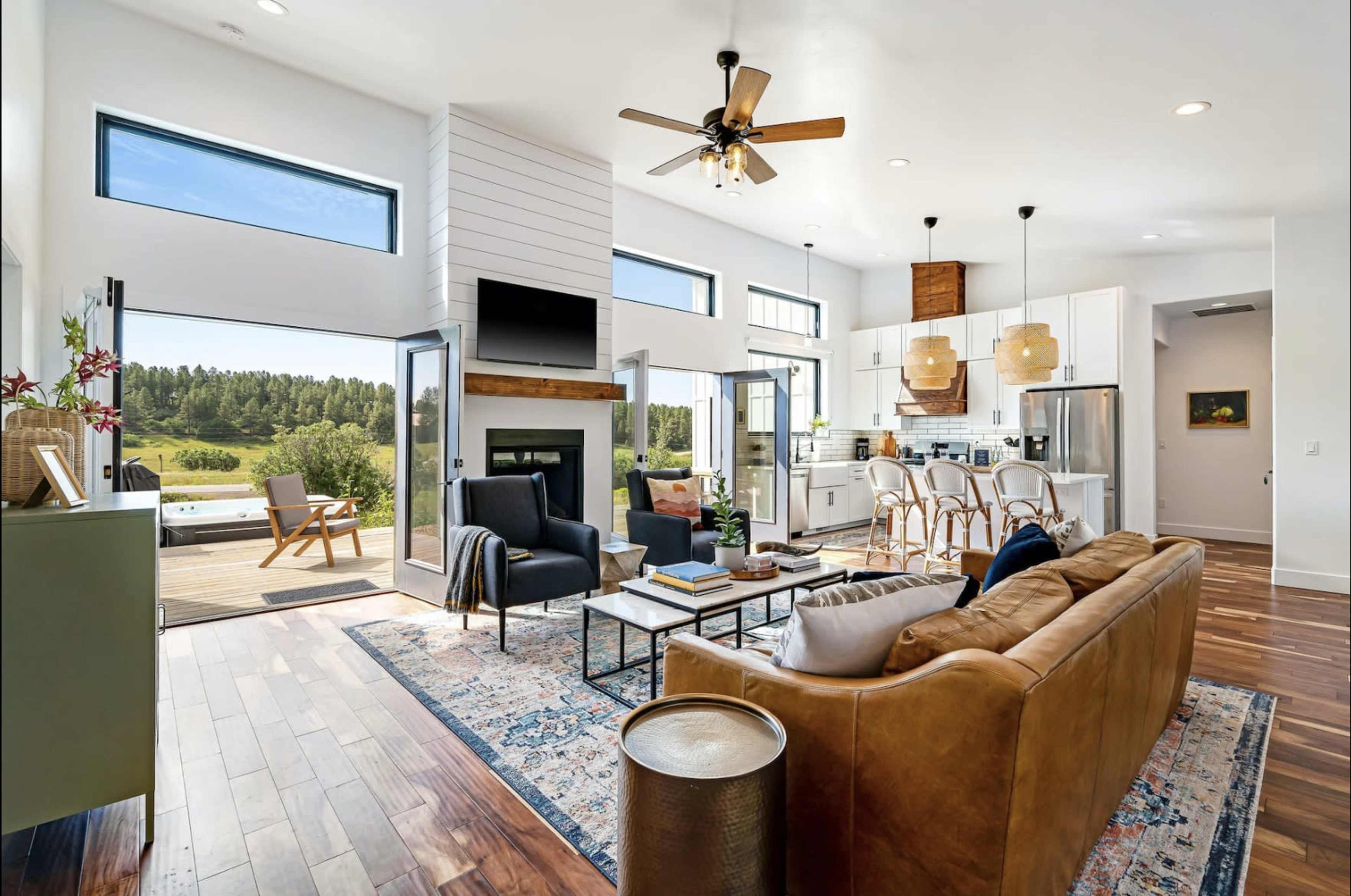 A modern living room with large windows and an open floor plan that connects to a kitchen area, featuring a brown leather sofa, wooden flooring, and a view of greenery outside.