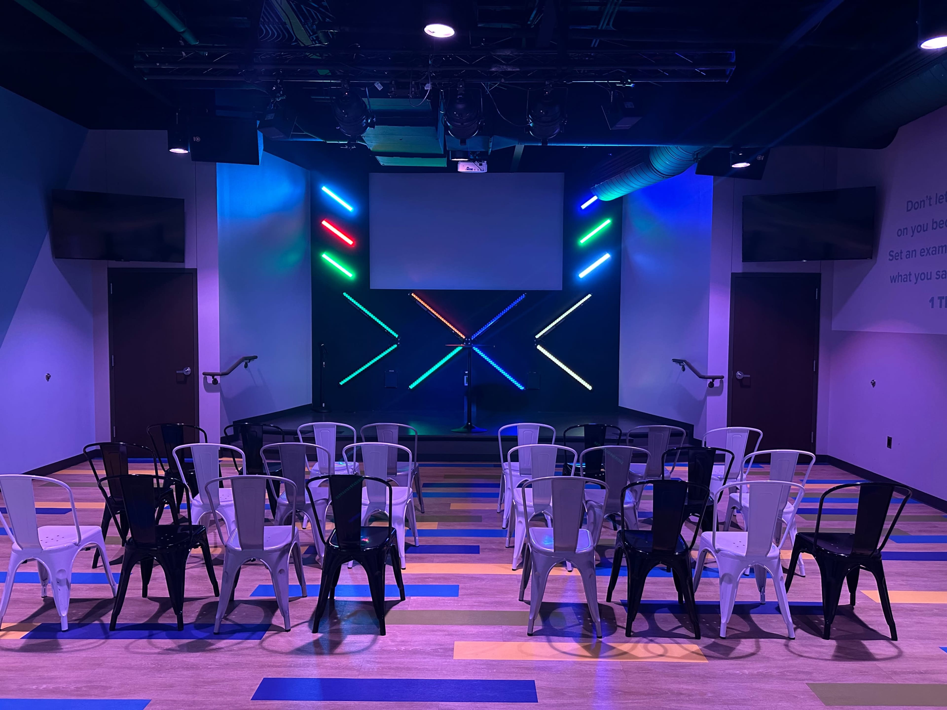 The image shows a brightly lit stage with colored lights and a row of folding chairs arranged in front of it in a modern indoor venue.
