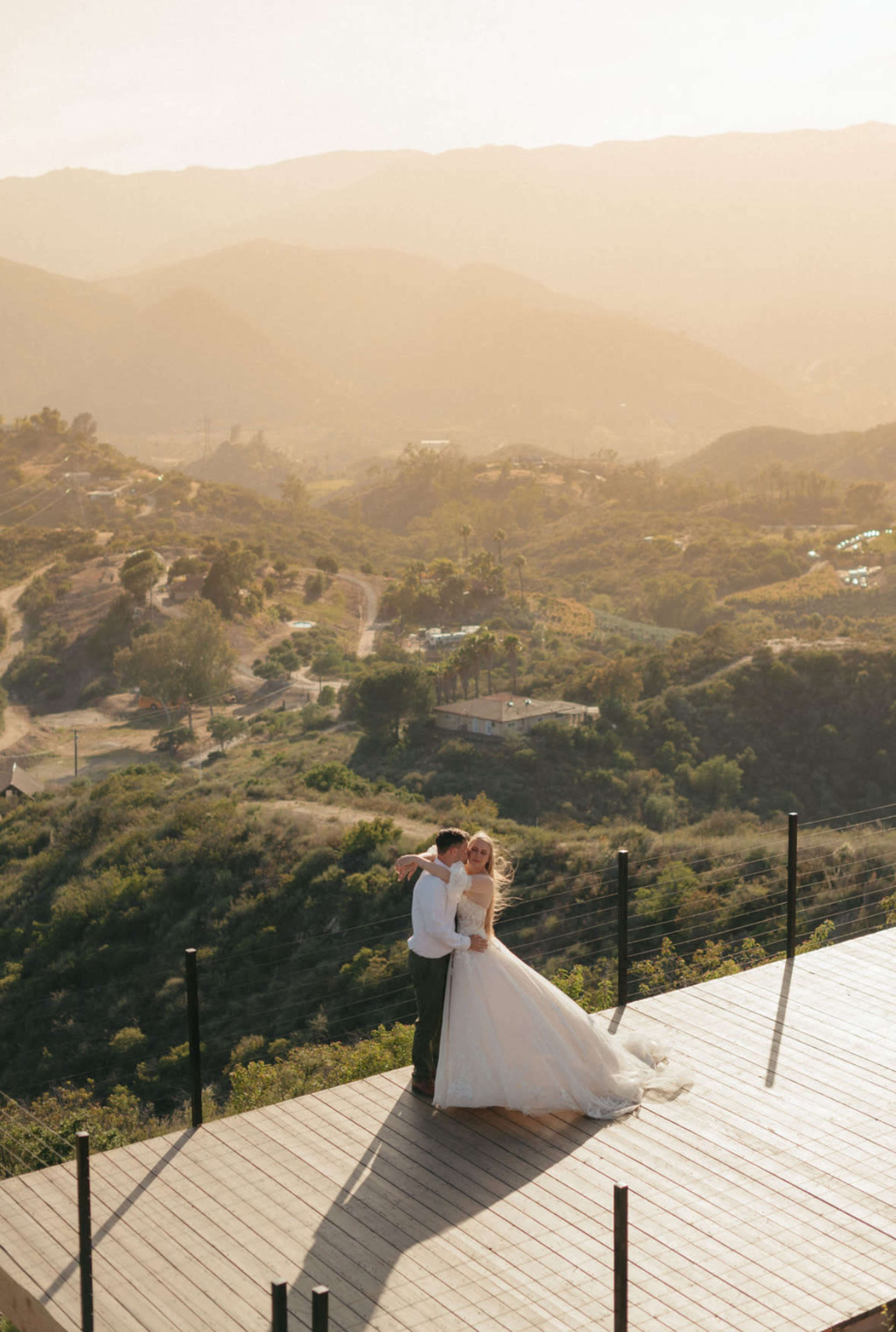 A bride and groom embrace on a wooden deck overlooking a mountainous landscape during sunset.