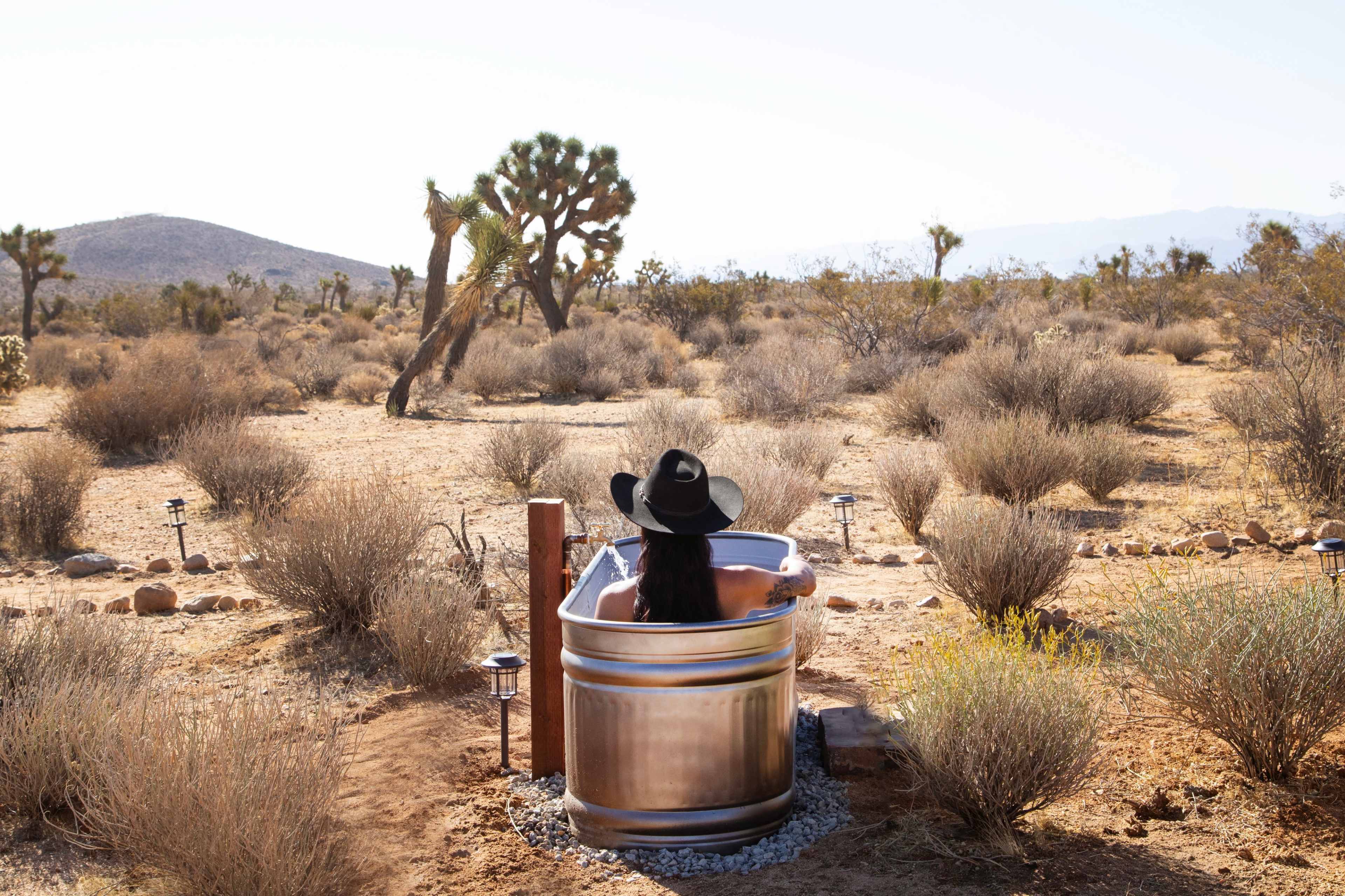 A person wearing a black hat sits in a metal bathtub situated in a desert landscape surrounded by sparse vegetation and distant mountains.