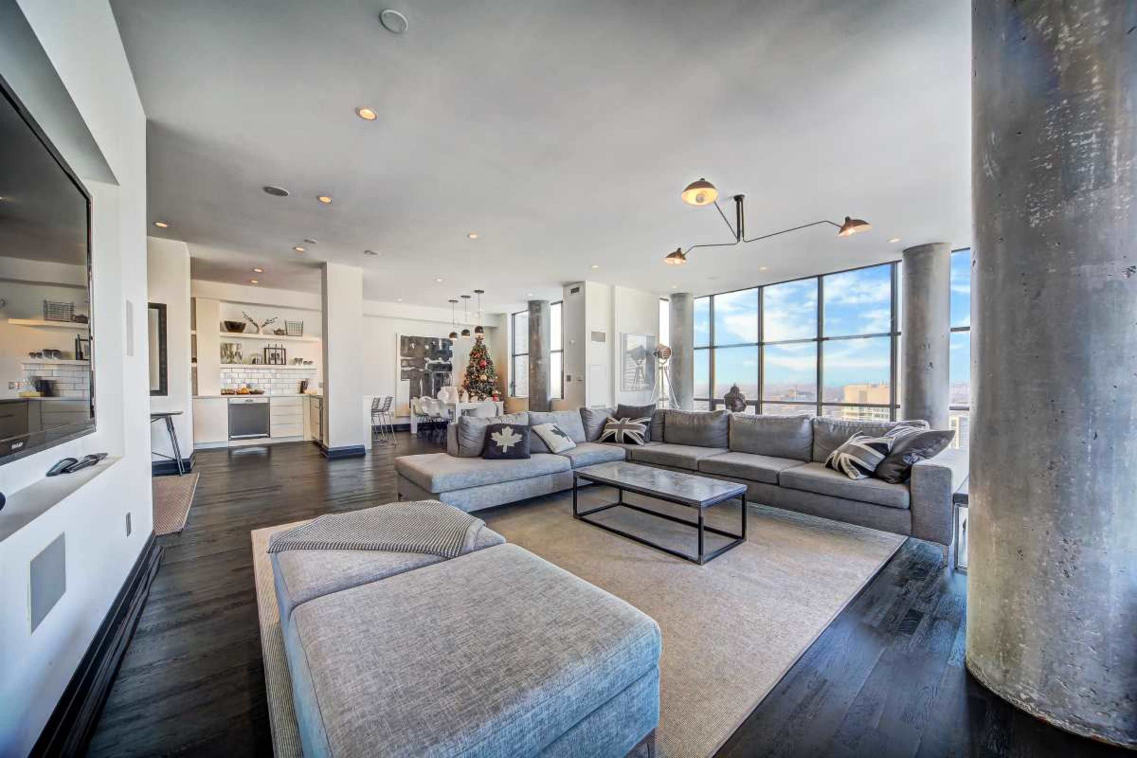The image shows a modern living room with large windows, a sectional sofa, and a coffee table in a high-rise apartment.