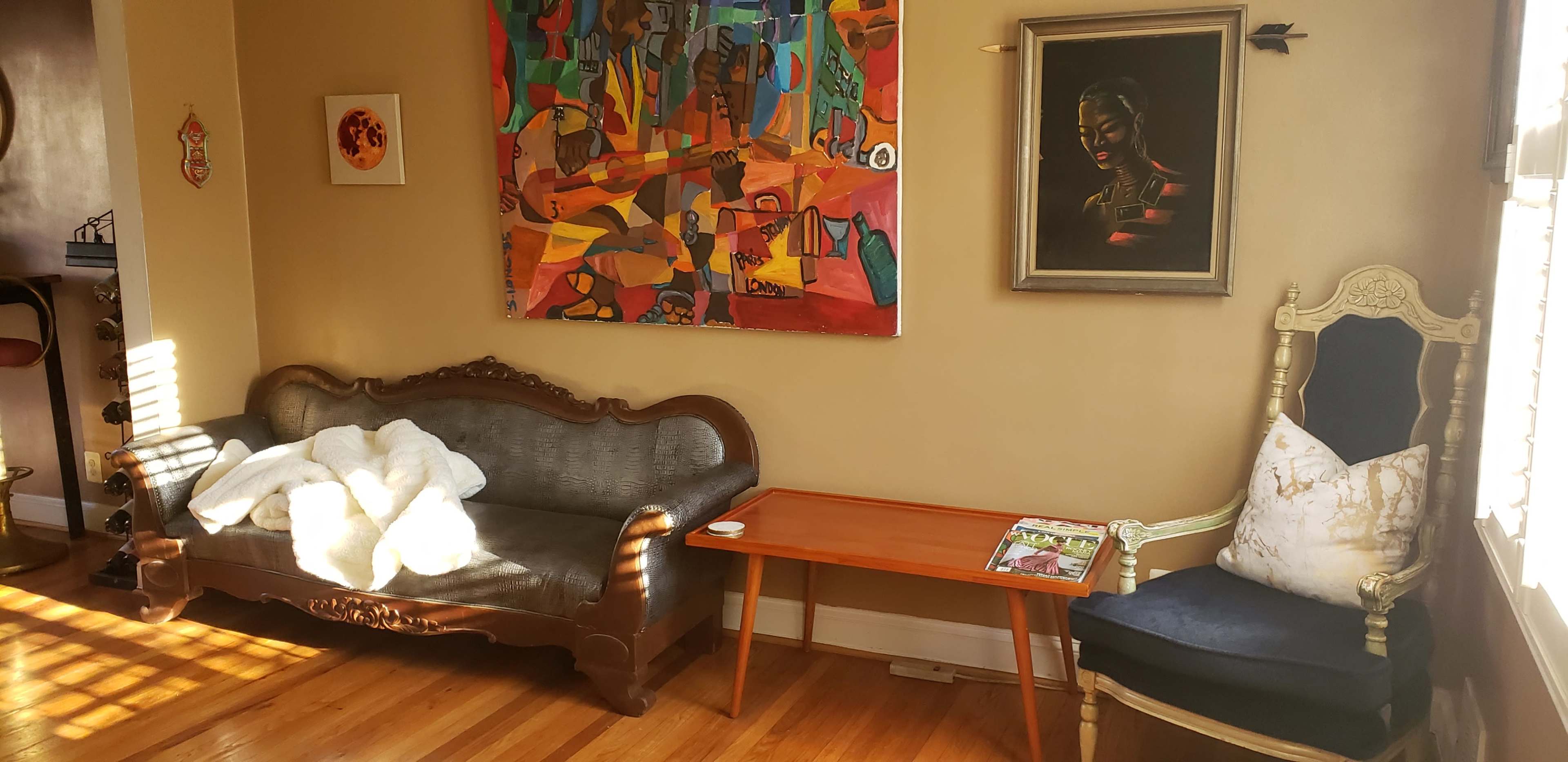 A living room featuring a dark sofa, an orange abstract painting on the wall, a wooden side table with a magazine, and a decorative chair with a cushion.