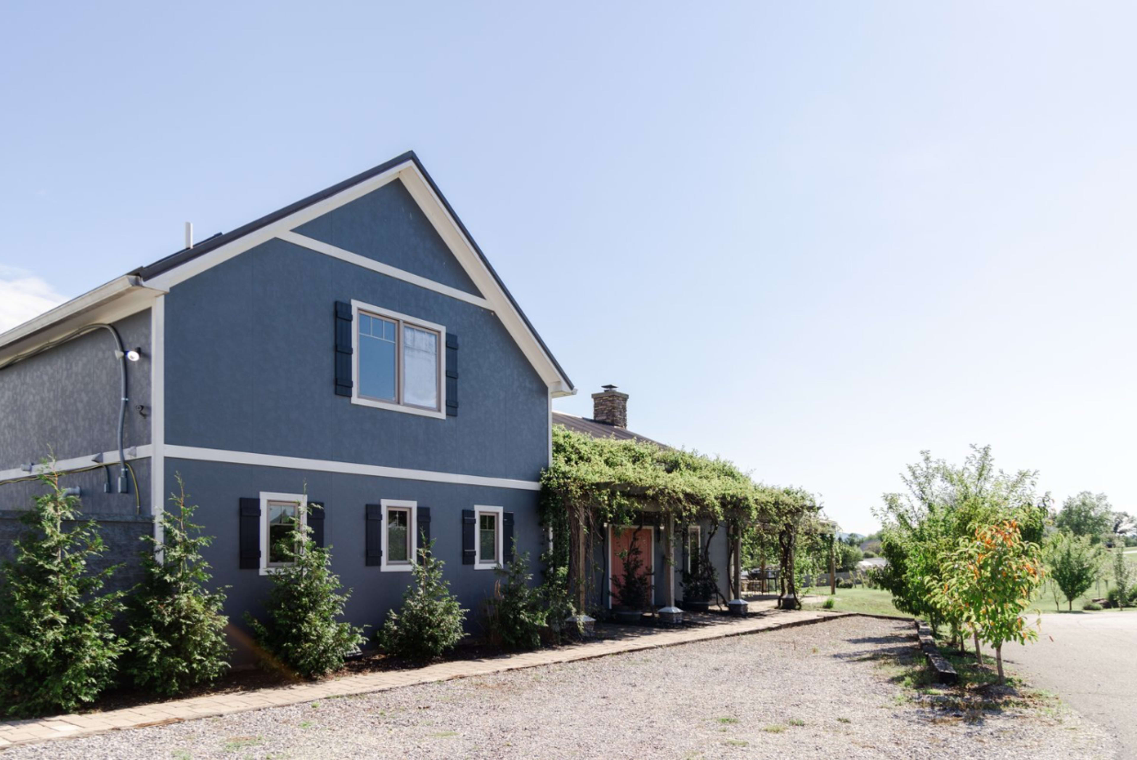 A blue two-story house with a covered porch and climbing vines is shown beside a gravel driveway and green landscape.