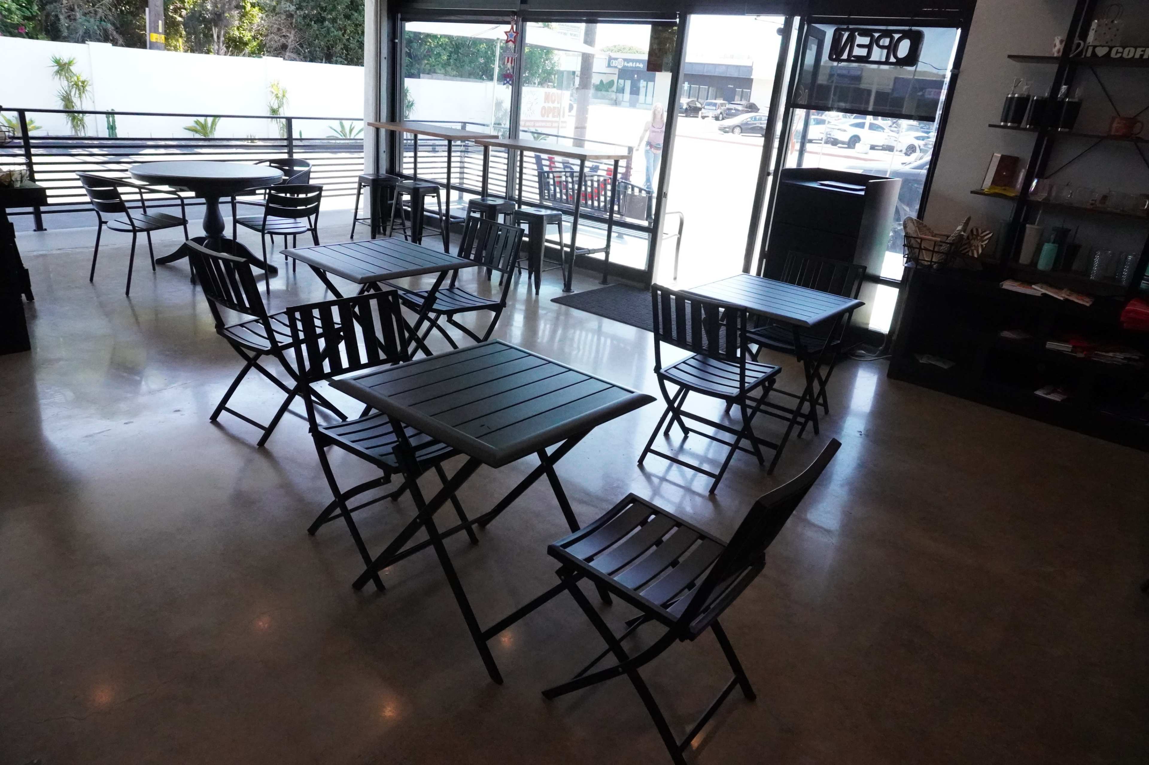 A cafe interior with several black metal folding tables and chairs arranged around a spacious area, featuring large windows that allow natural light.