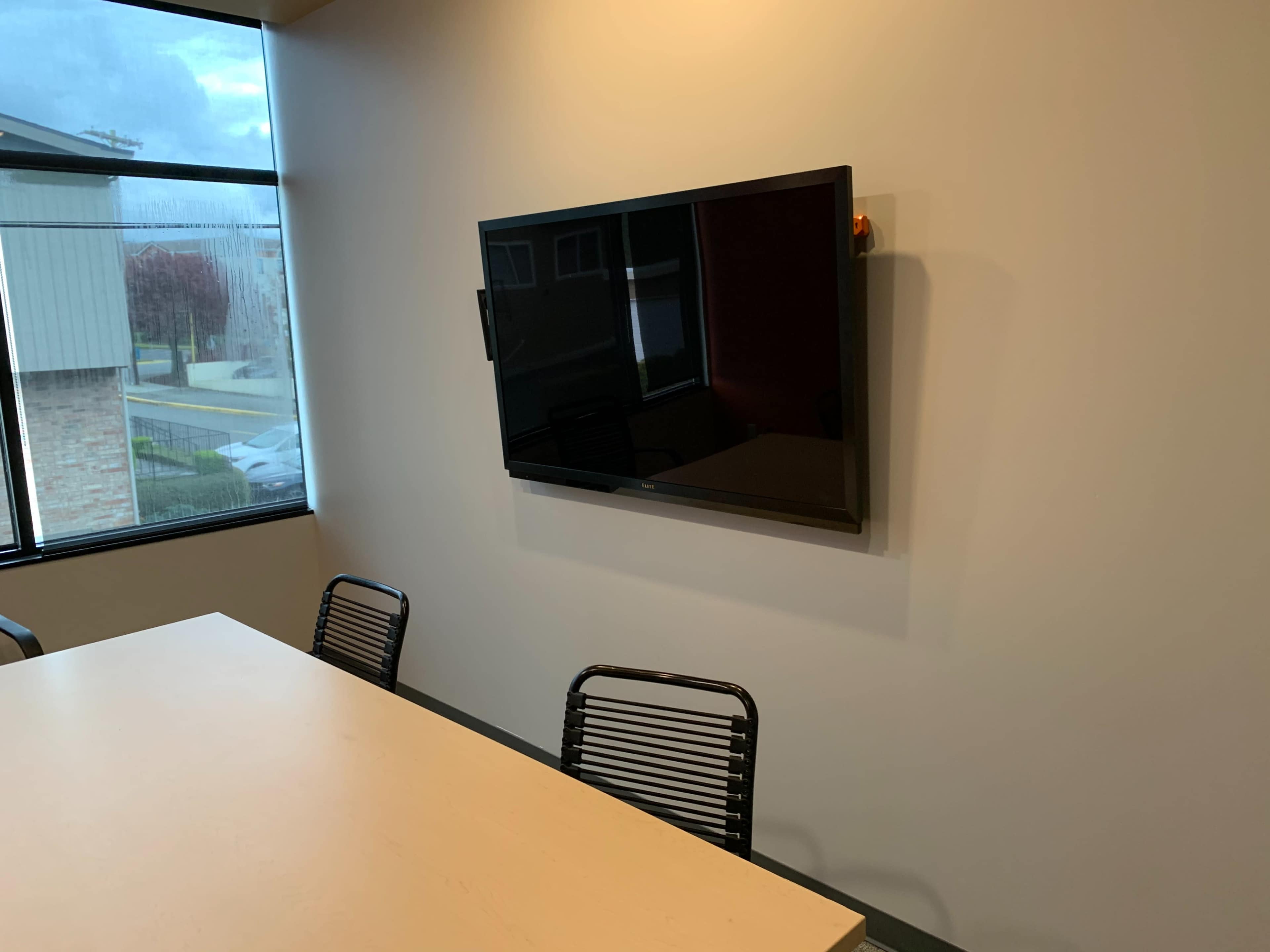 A mounted television on a light-colored wall in a meeting room with a table and chairs.