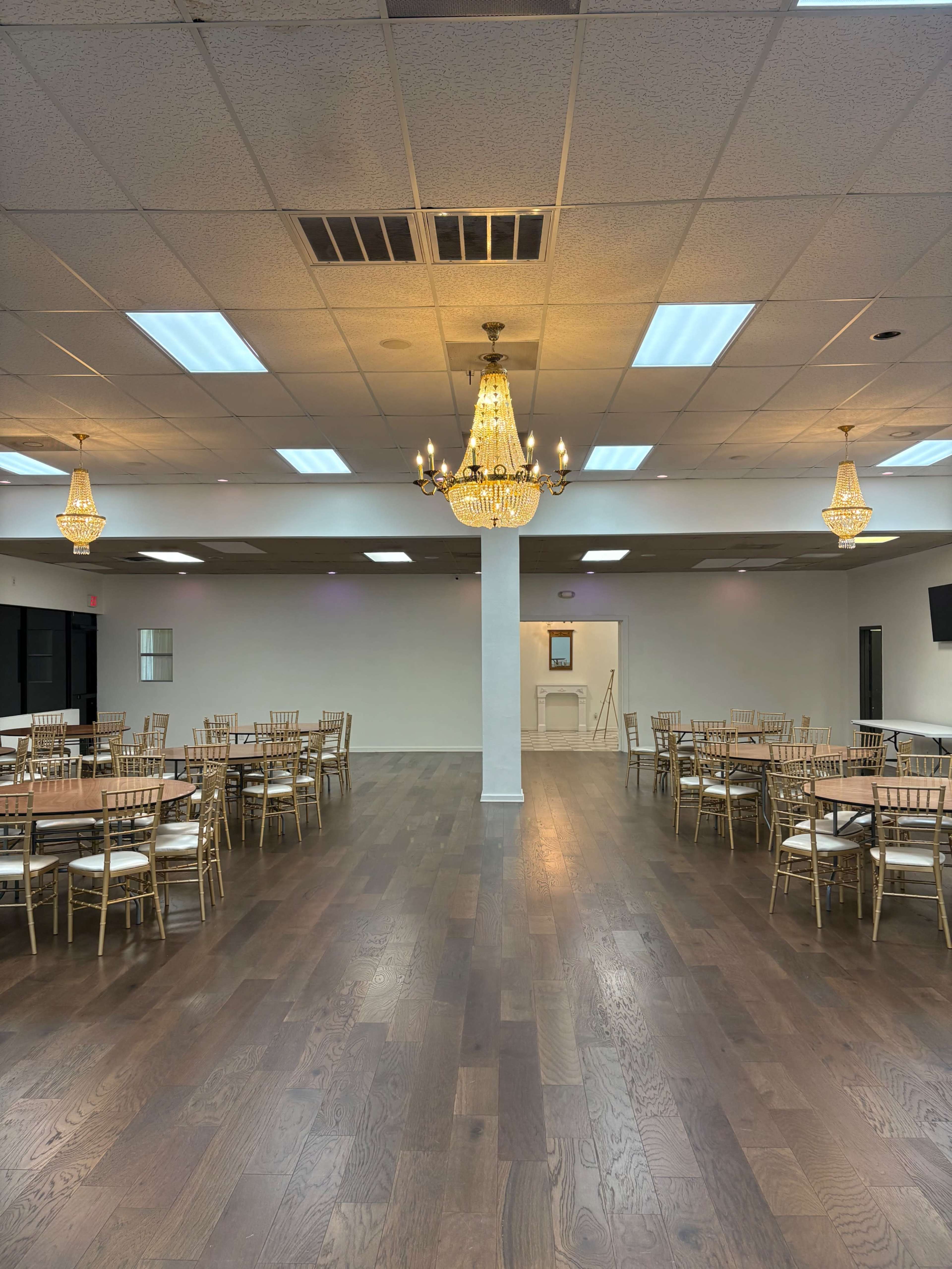 The image shows a large, empty banquet hall with wooden floors and several round tables arranged around a central chandelier.