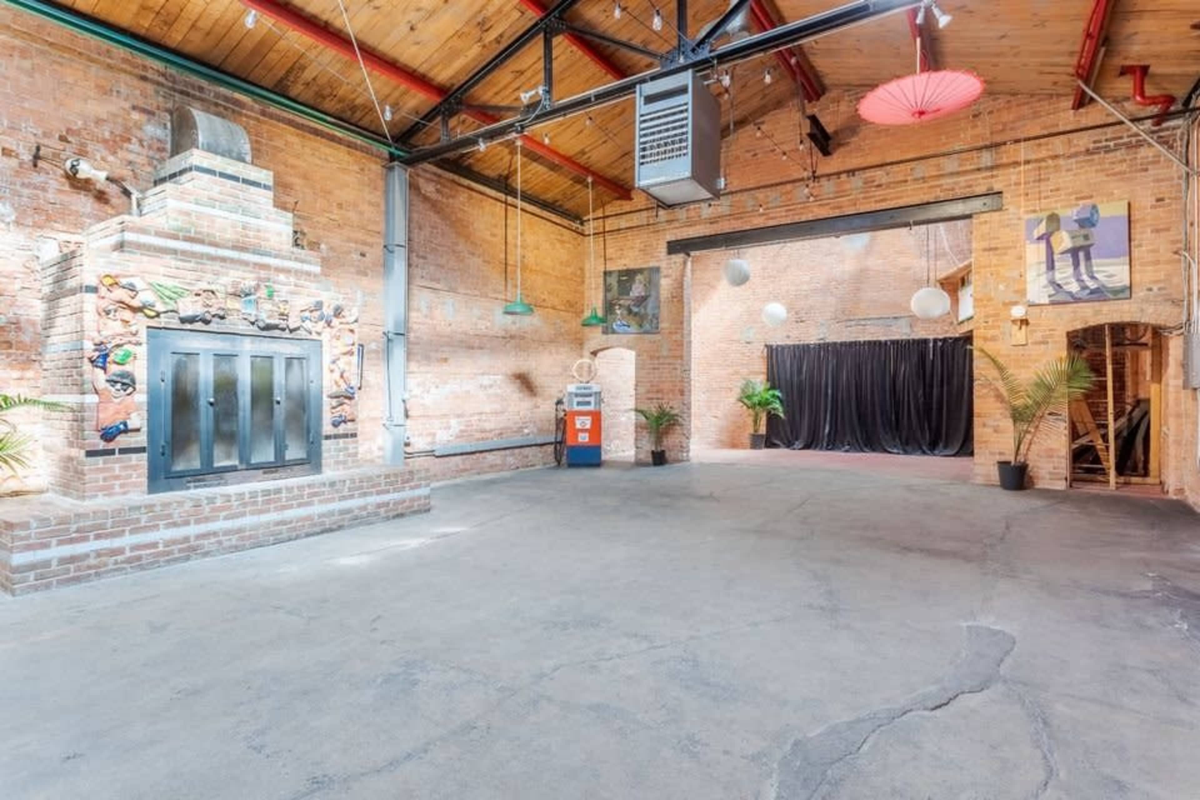The image shows a spacious, industrial-style interior with exposed brick walls, a concrete floor, and minimal furnishings, featuring a brick fireplace and decorative elements on the walls.