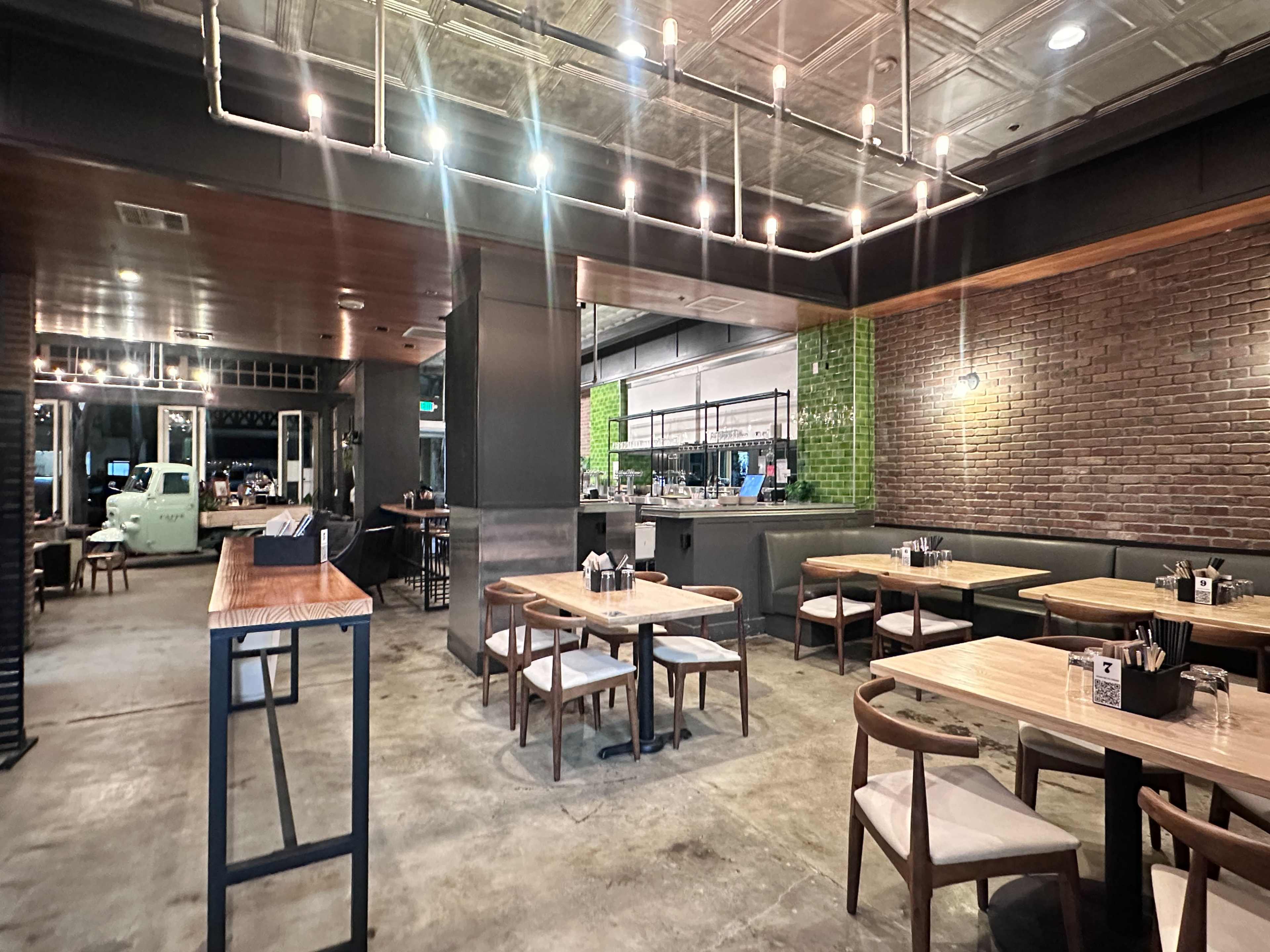 The image shows a modern restaurant interior featuring wooden tables and chairs, exposed brick walls, and industrial lighting.